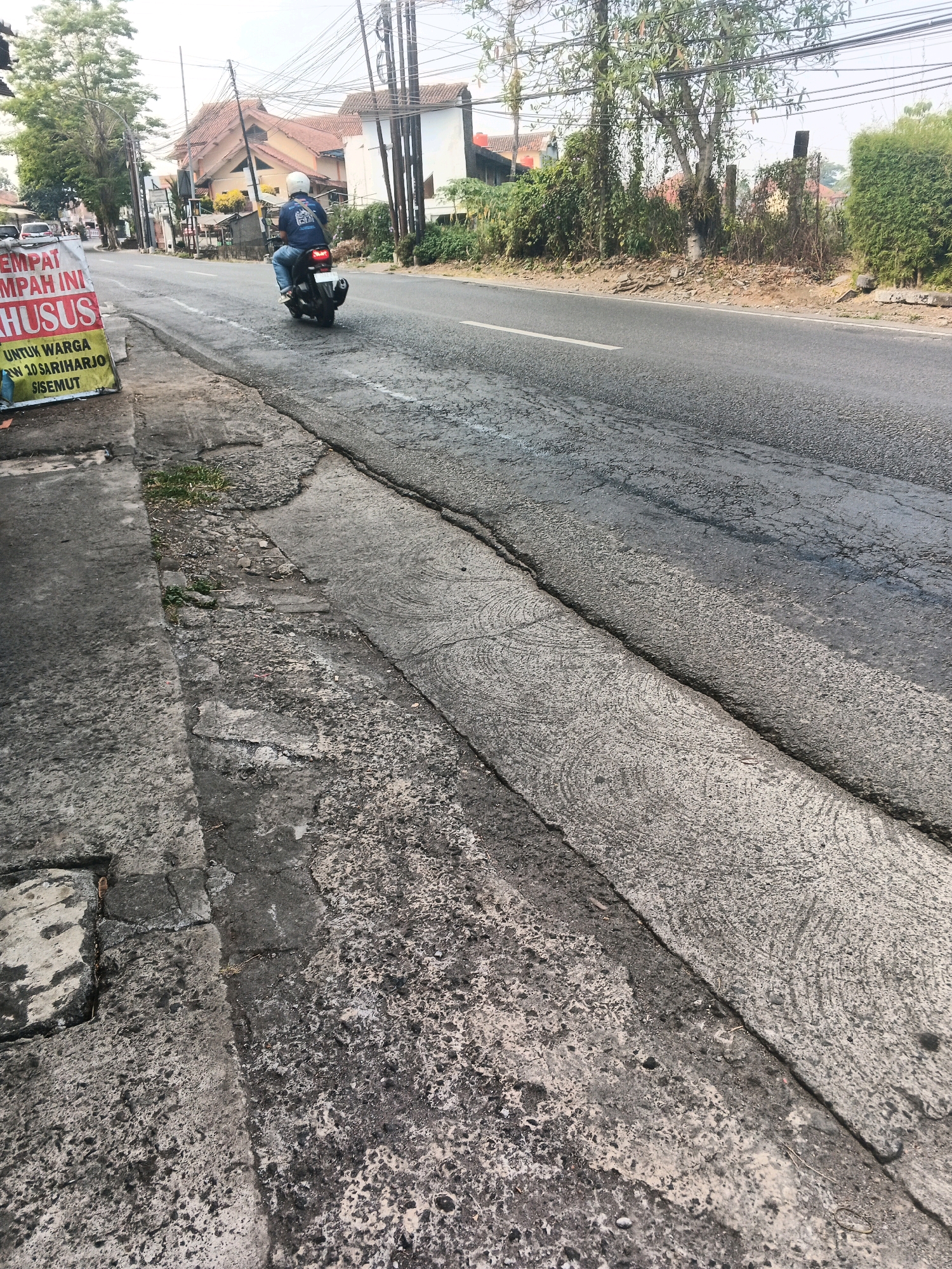 A person wearing a blue jacket and a white helmet rides a black motorcycle on a paved road in Ungaran, Indonesia. The motorcycle has a red taillight illuminated. To the left of the road, a sidewalk is visible, with a sign that reads "TEMPAT INI KHUSUS UNTUK WARGA RW 10 SARIHARJO SISEMUT". The road appears to be cracked in several places, and there are trees and buildings in the background. The sky is overcast.