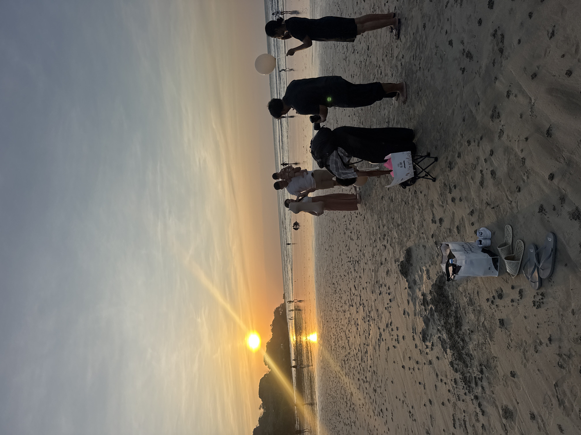 This image depicts a sunset scene on a beach in Kuta Selatan, Indonesia. The sun, a bright orange disc, is low on the horizon, casting a golden glow and reflecting on the wet sand near the water's edge. Silhouetted figures of people are scattered across the beach, some walking, some standing in groups. In the foreground, a group of individuals are gathered. One person is holding a camera, seemingly taking photos of another person who is holding a white balloon. Nearby, a small foldable chair is set up with a bag on it, and several pairs of flip-flops and shoes are arranged on the sand. The ocean stretches out in the background, with gentle waves rolling onto the shore. The sky is a gradient of pale blue and orange, with wispy clouds. The beach itself is sandy, with small mounds of sand and scattered debris. The overall atmosphere is tranquil and suggestive of a casual gathering or outing at sunset.
