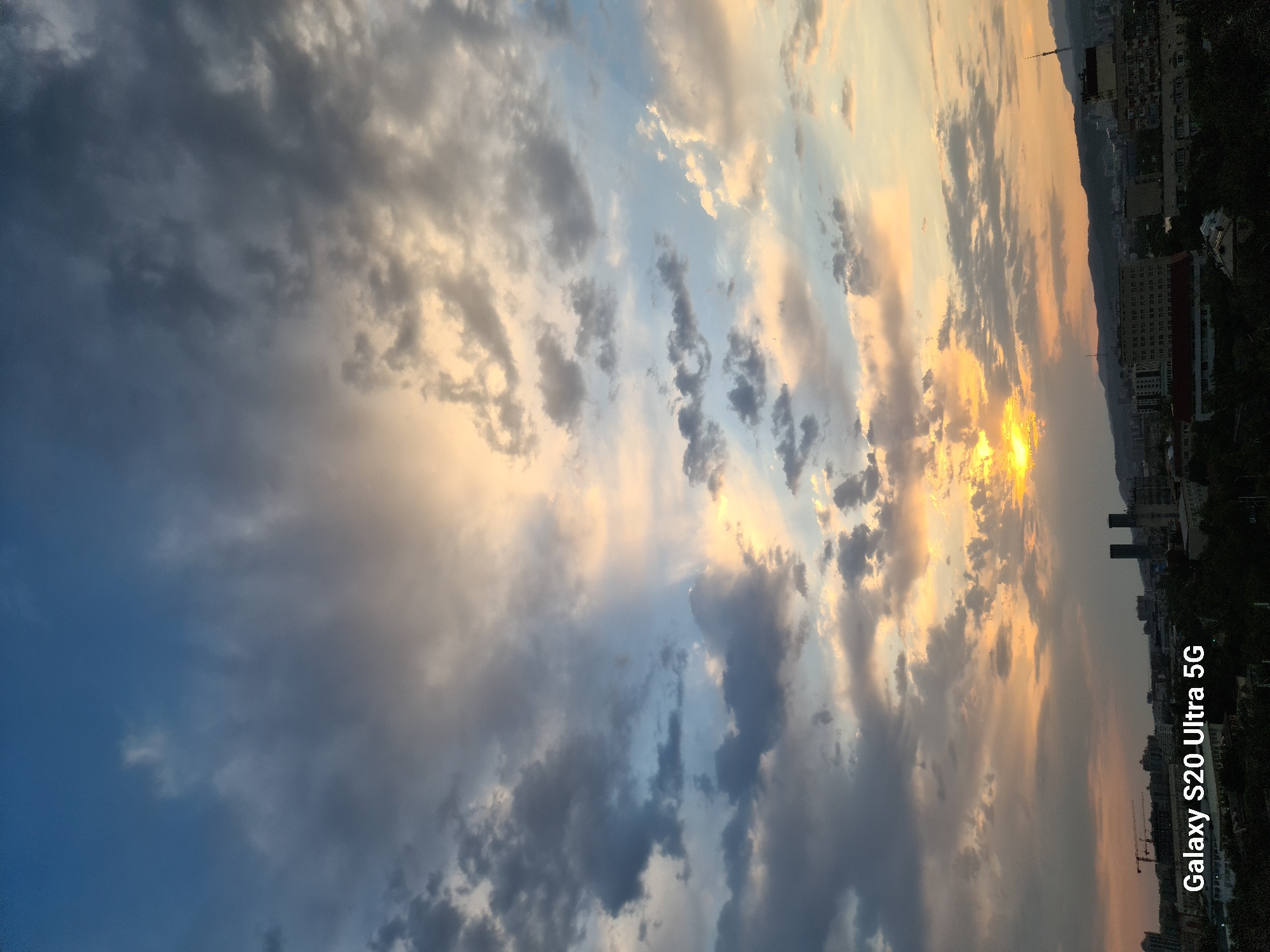 The image is a vertical, wide-angle photograph of a sunset over Almaty, Kazakhstan. The sky is filled with dramatic clouds, with streaks of orange and yellow light from the setting sun breaking through. Some clouds are dark and heavy, while others are lighter and wispy. In the foreground, a cityscape is visible, with buildings of varying heights, including some taller structures with smokestacks. Hills or mountains can be seen in the distance. A watermark "Galaxy S20 Ultra 5G" is present in the bottom left corner of the image. The overall impression is one of a dynamic and colorful natural phenomenon over an urban environment.