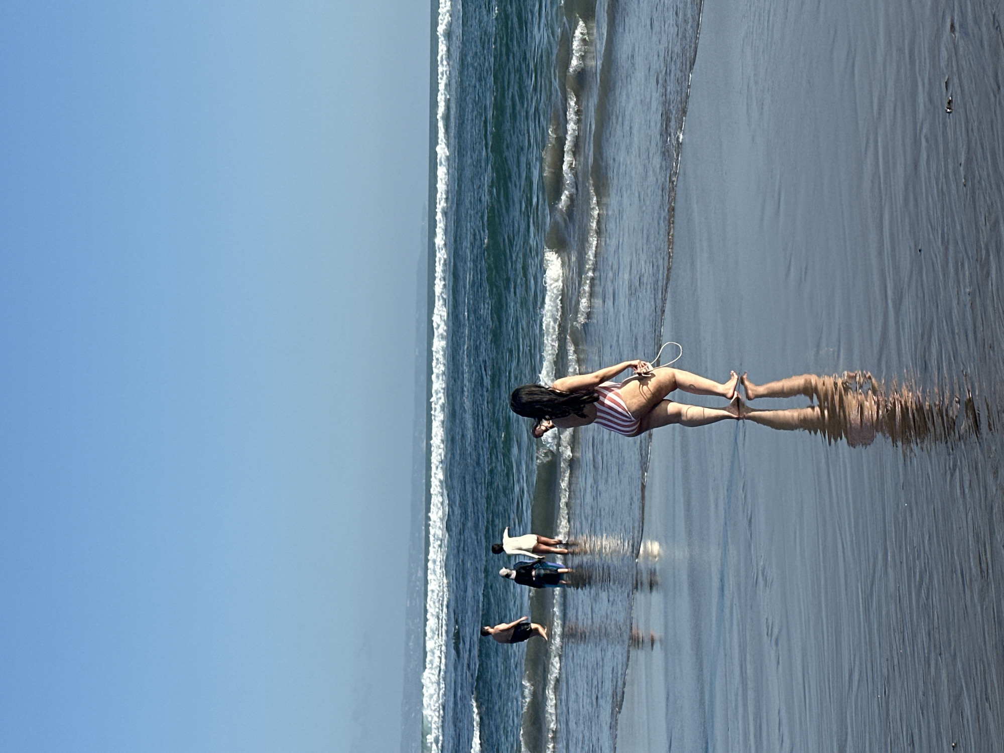 An outdoor, eye-level, vertical shot captures a beach scene on a clear day. The ocean, with moderate waves, fills the right two-thirds of the frame, extending to the horizon where land is visible. The foreground is a wet, reflective sandy beach. In the middle ground, four individuals are standing in the shallow ocean water. Two are men, one in dark shorts, the other in lighter shorts. Two are women, one in a white swimsuit and a cap, the other in a dark swimsuit. In the right foreground, a woman with long dark hair stands facing away from the camera, wearing a red and white striped one-piece swimsuit. Her reflection is visible on the wet sand. The sky is a clear, bright blue. This scene is characteristic of a beach location in North Kuta, Indonesia, such as Seminyak or Canggu, known for their shores, ocean access, and recreational activities.