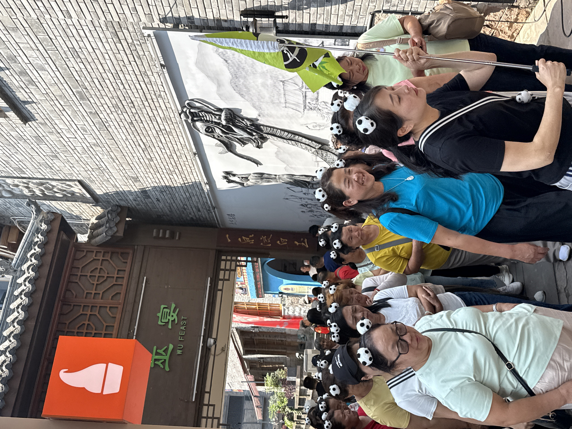 This image depicts a bustling street scene in Yuzhong District, China, under bright, sunny daylight. A large group of people, predominantly adults, are visible, many of whom are wearing distinctive black and white spherical clips, resembling miniature pandas or soccer balls, in their hair. This shared accessory suggests they might be part of an organized tour group or participating in a specific event.
The individuals are arranged in a line, seemingly queuing or moving through the street. Some are seen holding poles, possibly selfie sticks or tour guide flags, including a green flag with a black symbol. Many people in the foreground and midground are smiling or looking around, conveying a sense of enjoyment and engagement.
The setting features traditional Chinese architecture, including buildings with intricate wooden window patterns, tiled roofs, and brick walls. In the background, a large mural or banner displays a stylized dragon against a mountainous landscape. To the left, an orange sign with a white graphic resembling an ice cream cone is visible. Adjacent to it, a building features a dark wooden facade with the words "WU FEAST" in English, along with corresponding Chinese characters, indicating a restaurant or culinary establishment. The scene captures a lively atmosphere, likely a popular tourist spot or cultural district.