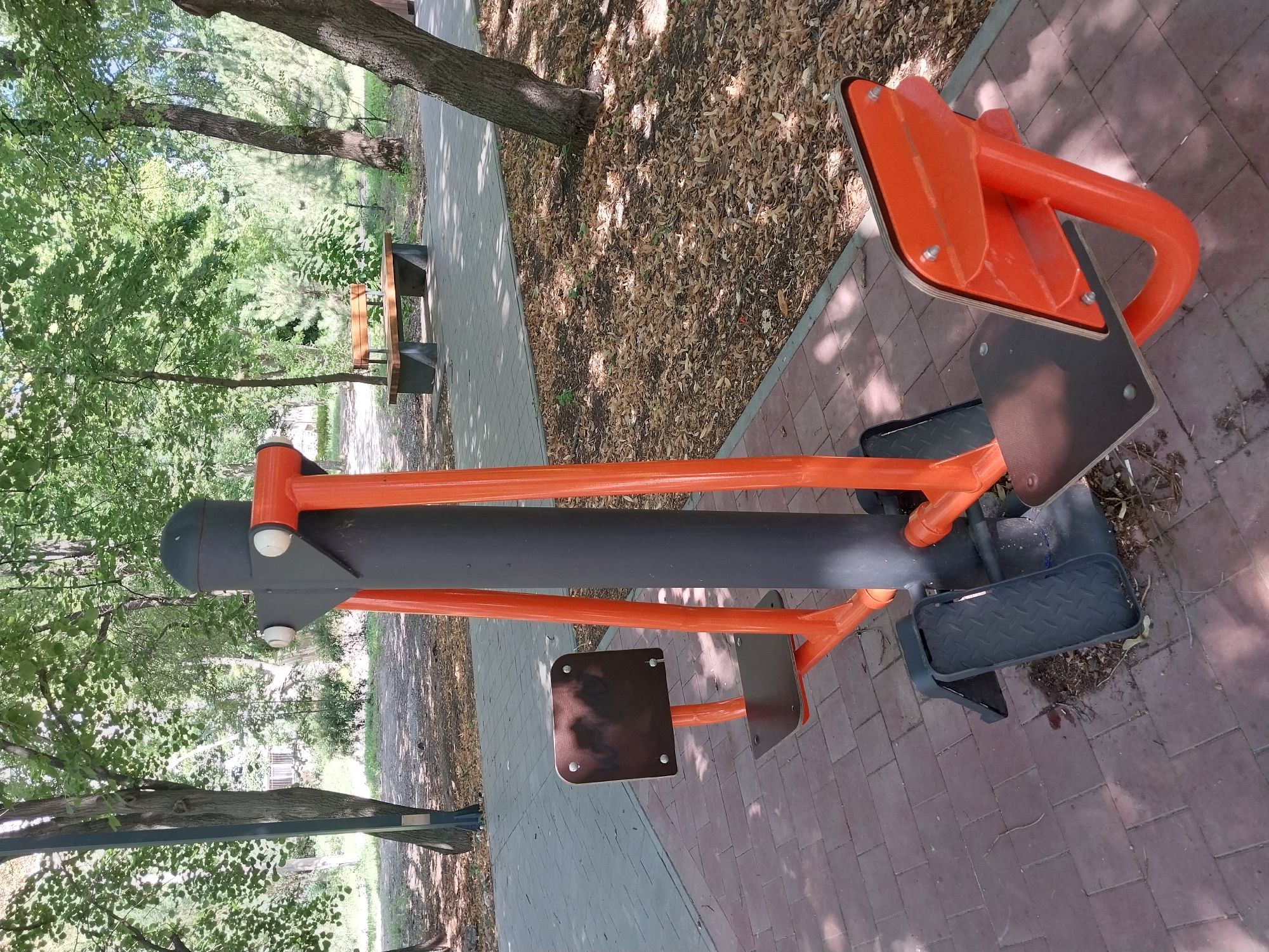 An outdoor fitness machine with orange and black metal components and wooden footrests and seats is positioned on a brick-paved walkway in a park. The machine appears to be an elliptical trainer. In the background, a wooden park bench sits on a paved area amidst trees with green foliage and fallen leaves scattered on the ground. The scene is bathed in dappled sunlight. This park is located in the city of Dnipro, Ukraine.