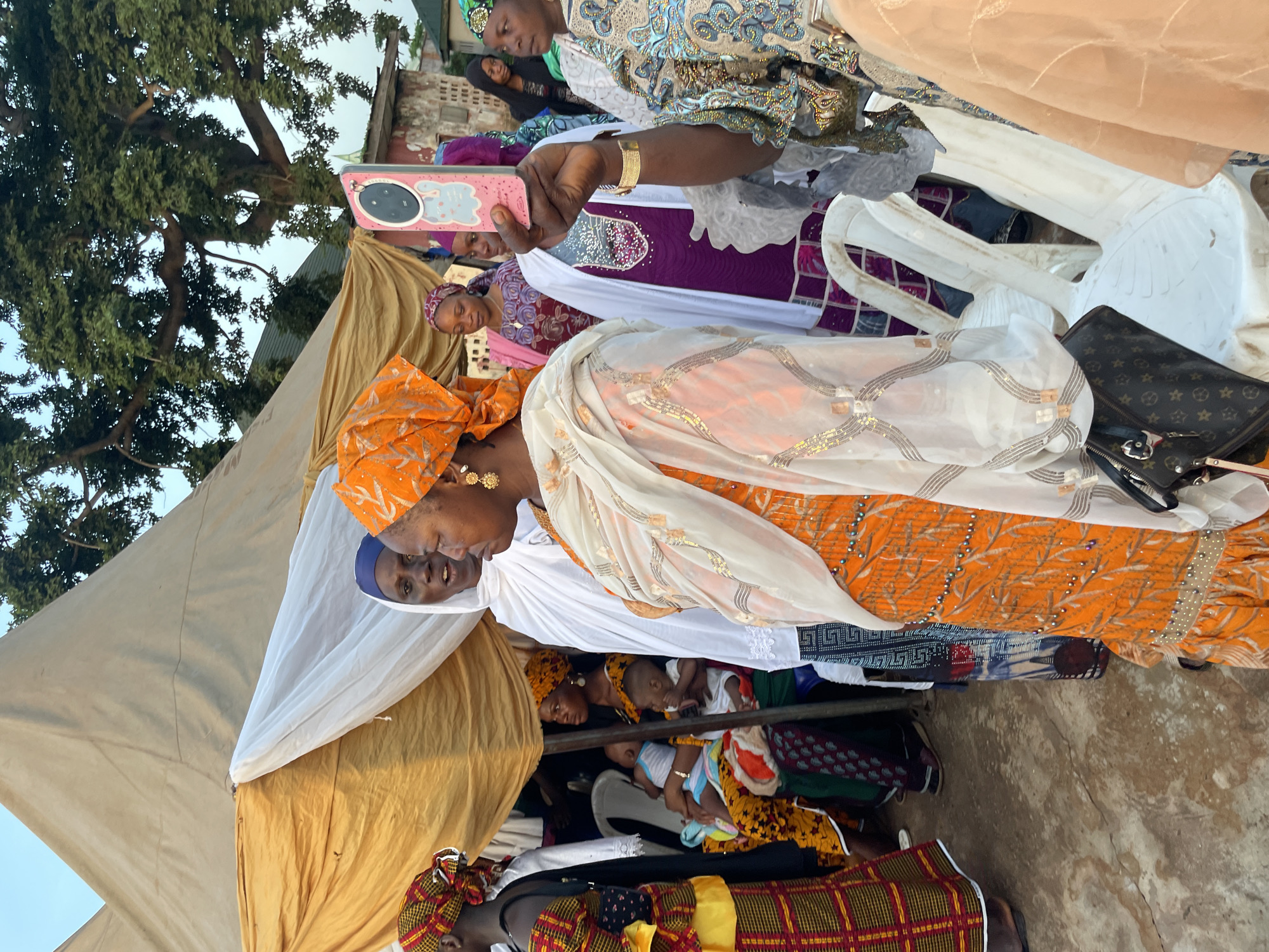 The image shows a gathering of people in what appears to be a community or outdoor event in Idah, Nigeria. Women are prominently featured, dressed in traditional attire with vibrant colors and headwraps. Some are holding babies or children. The location is outdoors, potentially in a residential area given the presence of buildings and a large tree. A tent-like structure with white and yellow fabric provides shade. An individual is taking a photo with a pink smartphone. There are white plastic chairs nearby, and a purse is visible on one of the chairs. The ground appears to be a mix of dirt and concrete.