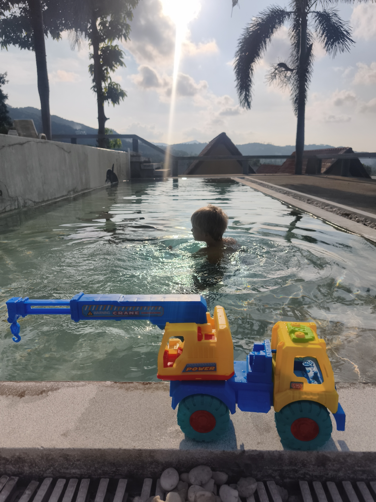 The image shows a child in a narrow, outdoor pool in what appears to be the city of บ้านบางรักษ์, Thailand. The child is swimming, with only their head and shoulders visible above the water. In the foreground, there is a large, plastic toy crane truck sitting on the edge of the pool. The crane is primarily yellow and blue, with red accents. The pool is surrounded by concrete edges. Beyond the pool, the background features trees (including a palm tree), hills, and possibly rooftops of structures. The sky is bright, with the sun shining through the scene.