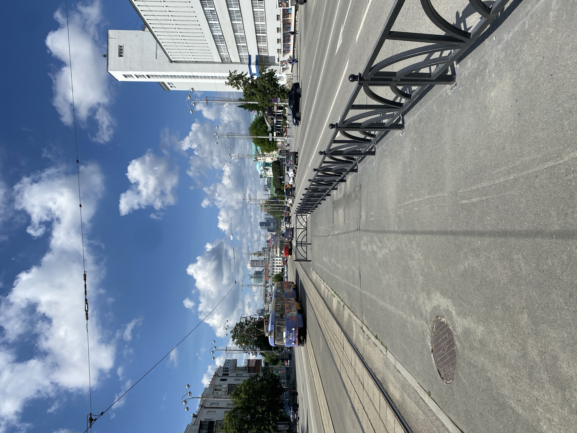 An outdoor, medium-shot, eye-level, vertical-format, full-color photograph captures a sunny day in Yekaterinburg, Russia. The image depicts a wide street with a tram line running along the left side. A blue tram with colorful markings is traveling on the tracks, with its pantograph connected to overhead wires. Several cars are visible on the multi-lane road, which is bordered by a sidewalk and a black metal fence. Buildings line the right side of the street, including a tall, white modernist structure. The sky is a vibrant blue with scattered white clouds. A manhole cover is visible on the sidewalk in the foreground. The overall scene conveys a typical urban streetscape.