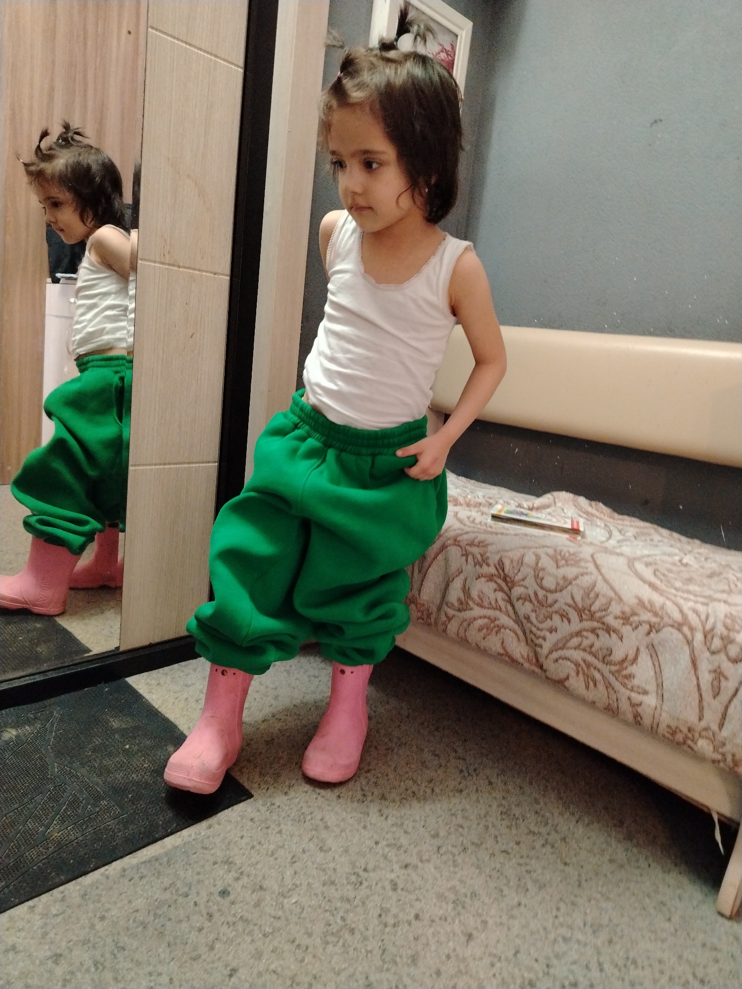 A young child stands in a room with a mirror. The child wears a white tank top, oversized green pants, and pink rubber boots. The child is leaning on a light beige bench with a patterned beige and brown cover. To the left of the child is a closet with a mirror. The child's reflection is visible in the mirror. A black mat is on the floor in front of the closet. The walls are gray. A framed picture is on the wall above the bench. A small book sits on the bench.