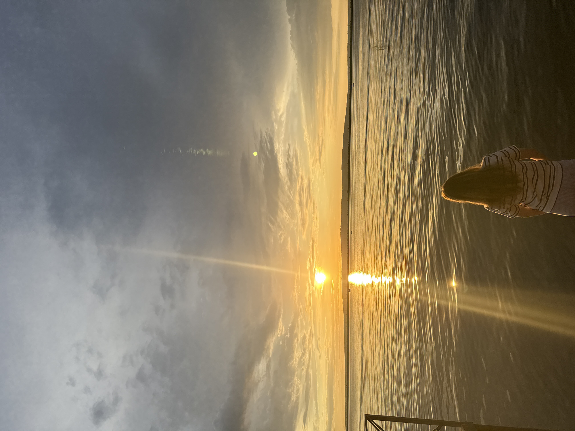 The image depicts a serene sunset over the Kama River in Chaikovsky, Russia. A young woman with long, light brown hair stands with her back to the camera, facing the water and the horizon. She is wearing a striped t-shirt and a light-colored skirt. The sun, a bright orange orb, is low in the sky, casting a brilliant golden reflection on the rippling surface of the river. The sky is a gradient of warm oranges and yellows near the horizon, transitioning to grayer, cloudier tones towards the top of the frame. In the distance, a faint silhouette of a treeline or distant buildings is visible along the riverbank. A small boat can be seen on the water, contributing to the peaceful ambiance of the scene. A metal railing of a pier is partially visible on the left side of the image. The overall impression is one of tranquility and natural beauty at the end of the day.
