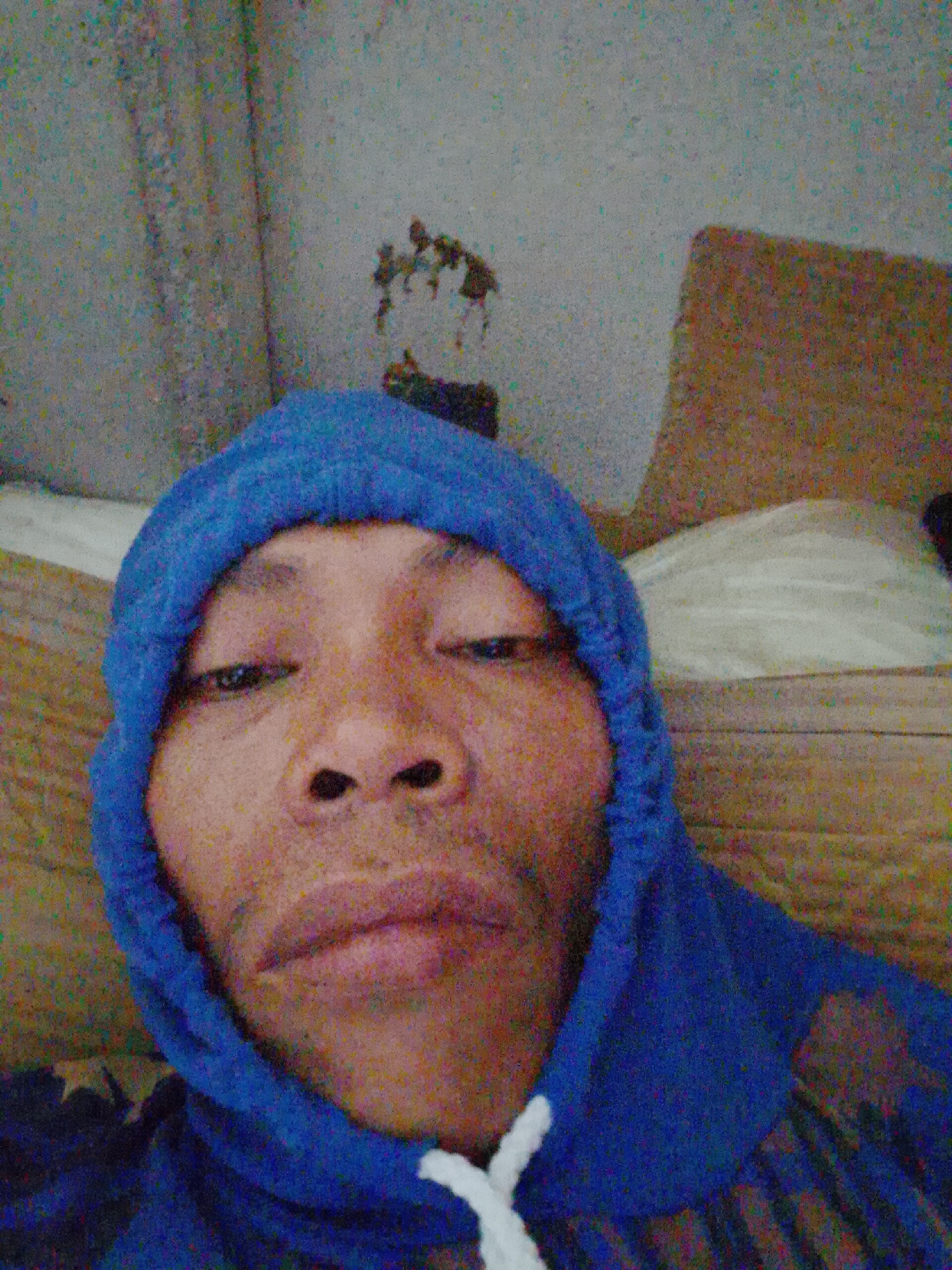 A close-up, indoor selfie shows a person with brown skin and dark eyes wearing a blue hoodie. The hood is pulled up, framing their face. The background consists of a wall with visible discoloration or markings, a white pillow, and a brown cushion. The location is unspecified in Bukit Ketok, Indonesia.