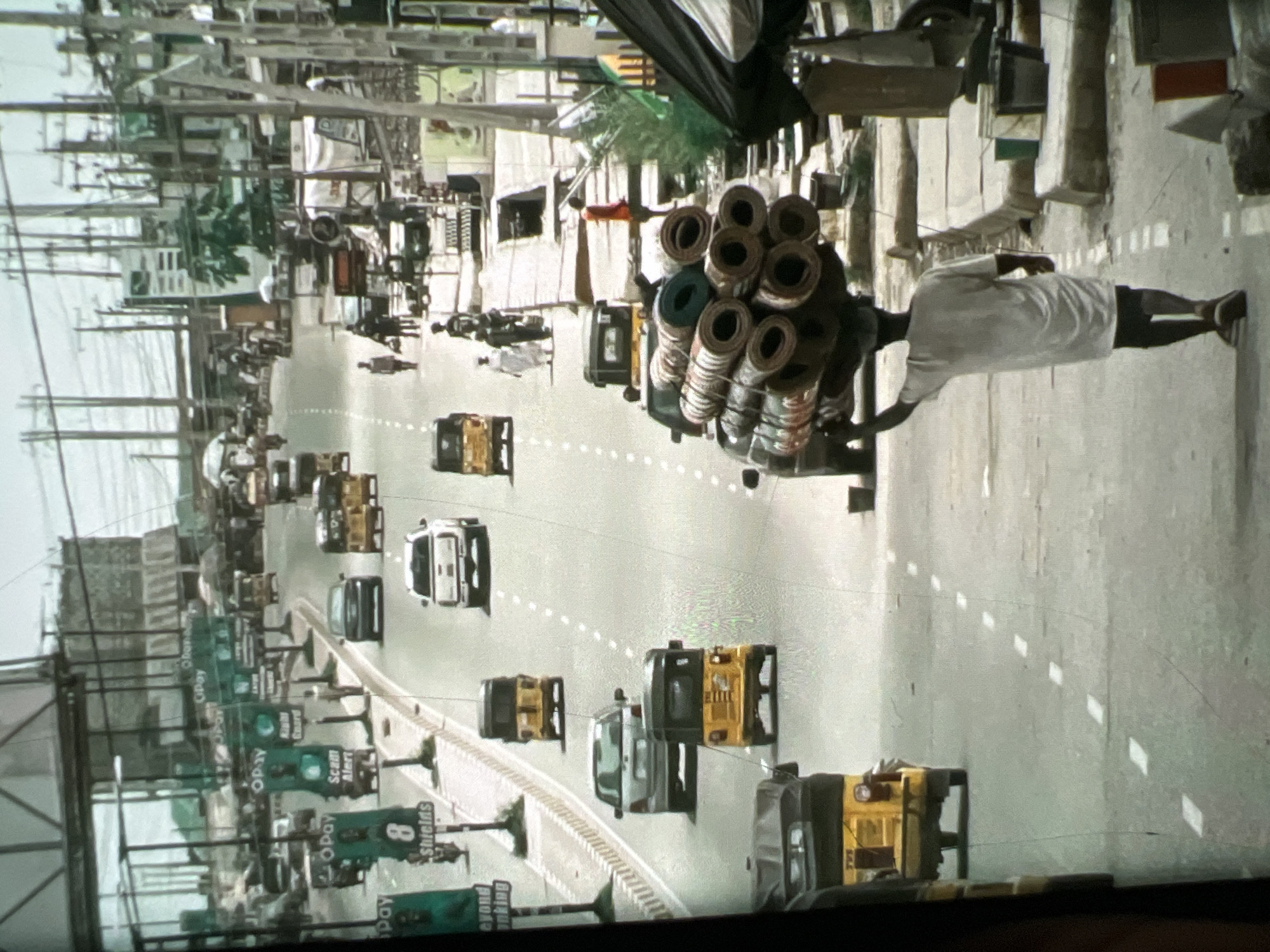 The image depicts a bustling street scene in Lagos, Nigeria, likely in a commercial or transportation hub. The foreground is dominated by a man walking away from the camera, carrying a large, circular load of rolled materials, possibly carpets or mats, balanced on his head. He is wearing a white t-shirt and dark shorts. To his left, a yellow auto-rickshaw, also known as a "Keke Napep" or "Marwa," is partially visible. Further down the street, several other auto-rickshaws and cars are navigating the road. The road is paved and marked with white dashed lines. Along the sides of the street, there are shops, market stalls, and numerous advertisements and billboards, many of which appear to be for financial services like OPay. Utility poles with a dense network of electrical wires are prominent in the background, suggesting an urban environment. The architecture is a mix of modern structures and older buildings, consistent with the varied development found in Lagos. The overall impression is one of a vibrant and active city with considerable pedestrian and vehicular traffic.