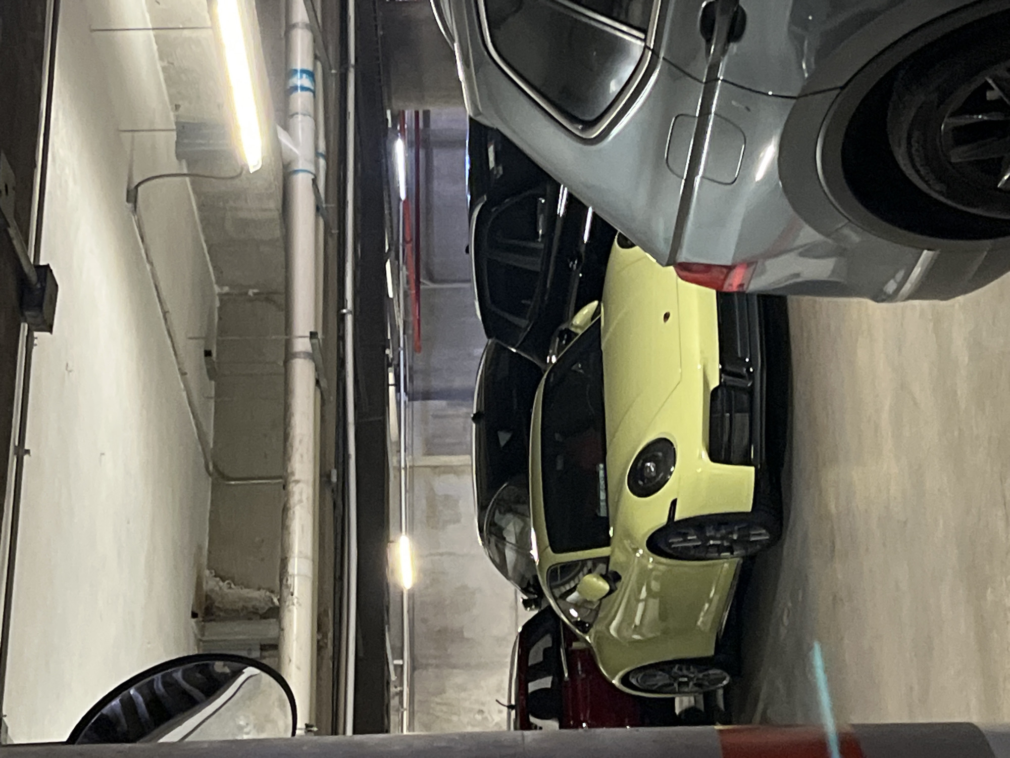 The image, taken in an indoor parking garage and rotated 90 degrees counter-clockwise from its natural orientation, displays several parked cars. In the immediate foreground, a striking light-yellow (or lime green) sports car, identifiable by its distinct rounded headlights and sleek design (likely a Porsche), is parked. Above it, a grey SUV or larger car is visible, its rear quarter panel, wheel, and part of a red taillight showing. Further back and slightly to the right of the yellow sports car, a dark-colored car is partially visible, followed by a hint of a red vehicle behind it.
The setting is a typical parking garage, characterized by concrete walls and ceilings. The space is brightly illuminated by long, horizontal fluorescent lights attached to the ceiling. Exposed utility pipes, predominantly white with some blue banding or markings, run along the walls and ceiling. The bottom left of the frame shows the reflection of the concrete wall and pipes in what appears to be the side mirror of the vehicle from which the photograph was taken. No people are visible, and the cars are stationary, suggesting a mundane parking activity. There is no clearly legible text in the image; the blue markings on the pipes are indistinct. The overall scene suggests a well-maintained, possibly upscale, parking facility.