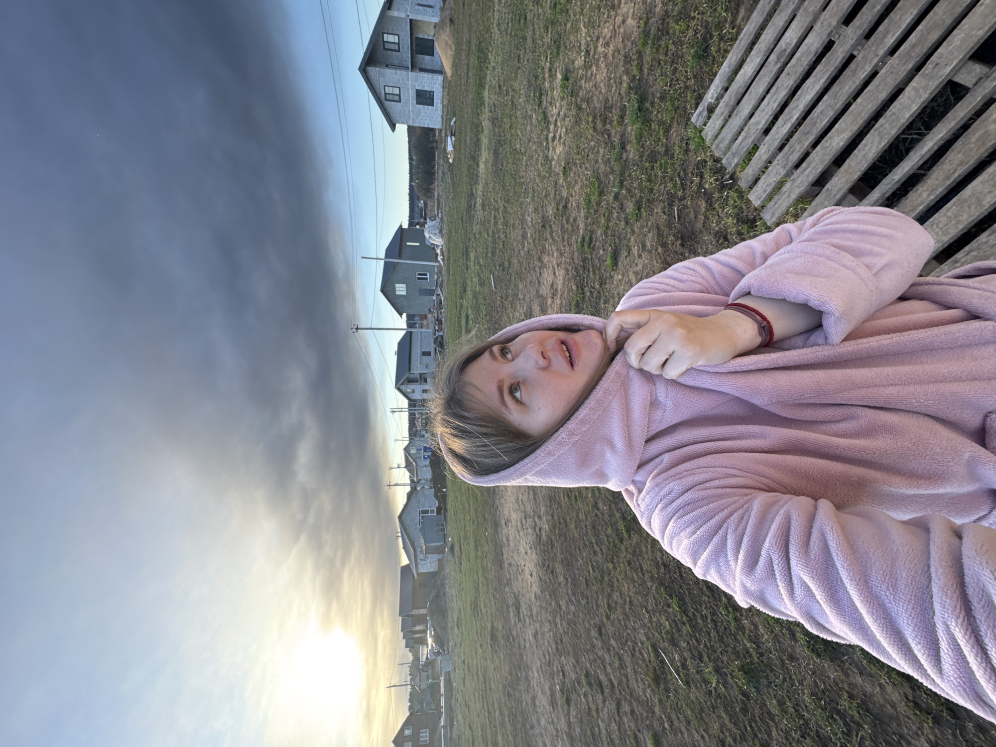 A young woman, wearing a pink hooded robe or towel, is in the foreground, appearing to be seated or leaning, and looking upwards towards the left with a neutral, observant expression. Her light brown hair frames her face, and a red string bracelet is visible on her right wrist.
The background depicts a semi-rural or developing residential area in Ферма, Russia. Several two-story houses, constructed with concrete blocks, are scattered across the mid-ground, some appearing to be newly built or under construction. The ground is a mixture of sparse green grass and bare earth. To the far right, a wooden slatted structure, possibly a bench or pallet, is visible.
The sky occupies a significant portion of the image, showing a transition from a bright, warm, low sun on the left horizon to a darker, more overcast cloud cover towards the upper right. This suggests the time of day is late afternoon or early evening. Utility poles and overhead power lines are visible, stretching across the scene. There is no visible text in the image.