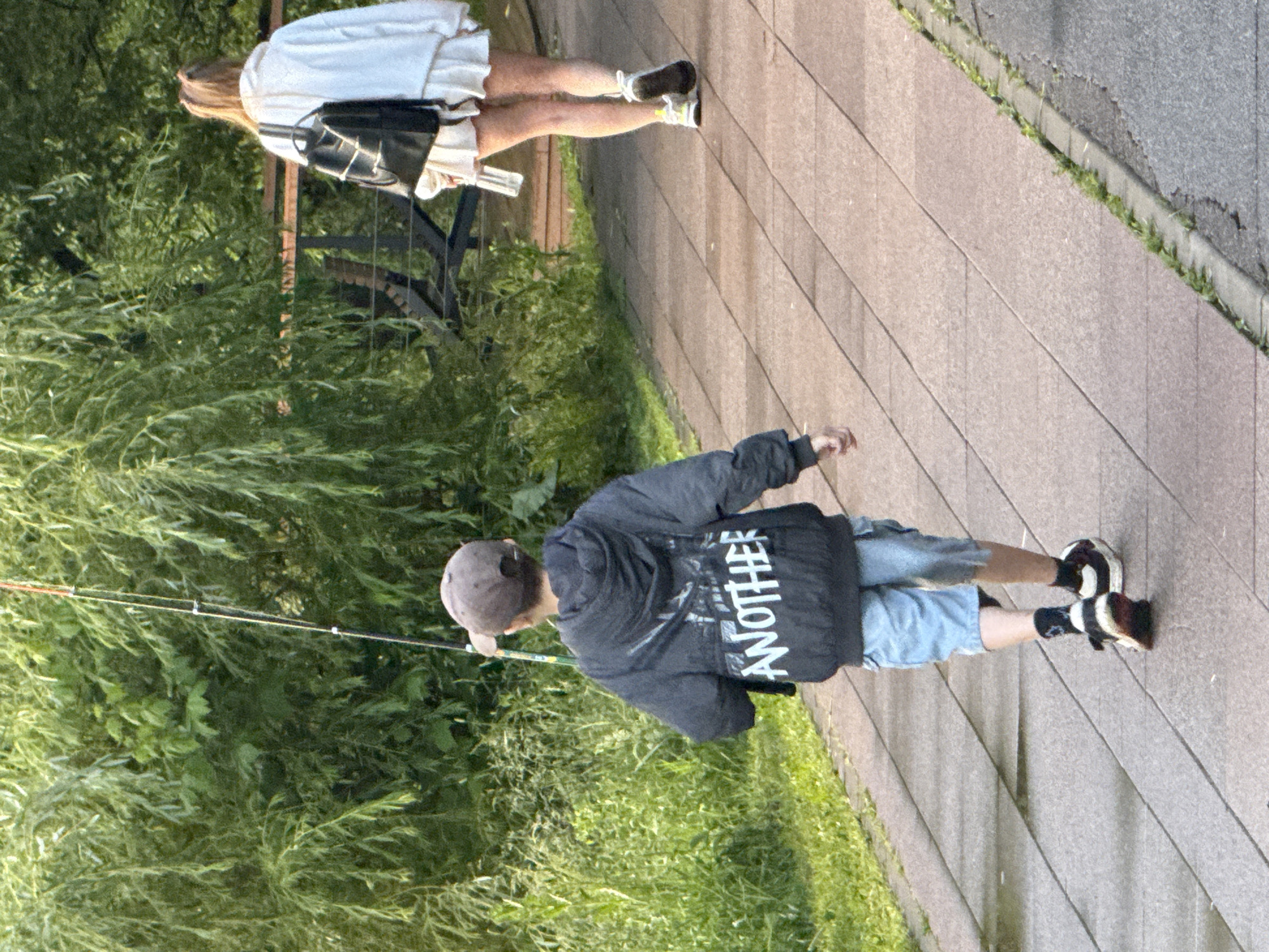 In Moscow, Russia, on a paved walkway lined with green grass and trees, a young boy walks away from the camera holding a fishing rod. He wears a dark gray hooded jacket with white lettering that reads "ANOTHER" on the back, light blue shorts, and black and white sneakers. Behind him, a young woman also walks away from the camera, wearing a white oversized hoodie and a white skirt. She carries a black backpack. The scene appears to be an outdoor recreational area.