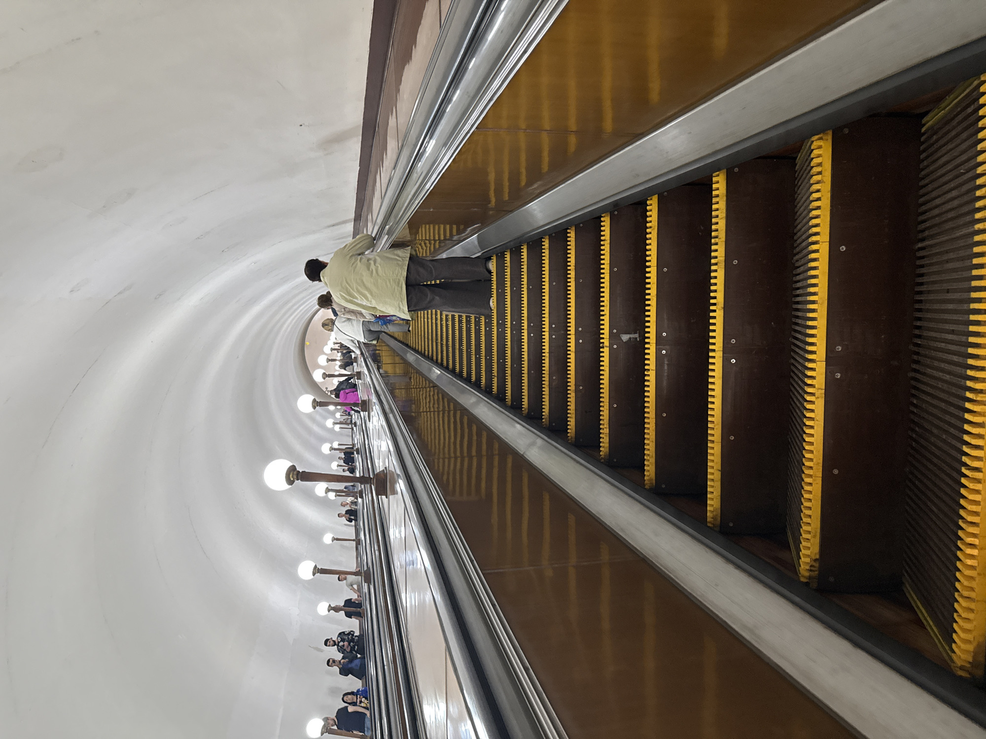 The image shows a long, arched escalator in a subway station, likely in Moscow, Russia, given the architecture. The escalator is well-lit by rows of spherical lamps mounted on tall, thin poles, spaced along the sides of the tunnel. The ceiling is a smooth, white, arched structure. Several people are visible ascending the escalator, some standing and some walking. The steps of the escalator are dark brown with bright yellow safety edges. The sides of the escalator appear to be made of polished, dark brown material. In the background, the escalator continues to ascend into the distance, suggesting a significant depth to the station. The perspective is from a low angle, looking up the escalator.