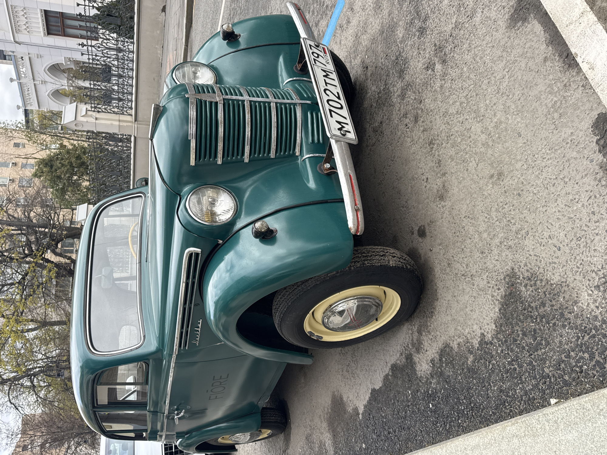 The image captures a vintage dark green car, likely a Moskvich (indicated by cursive text on the side, partially visible), parked on an asphalt street in Moscow, Russia. The car is viewed from a three-quarter front perspective, showcasing its classic design elements.
Key features of the car include a vertical chrome grille, prominent round headlamps, and a chrome bumper with a visible Russian license plate reading "M 702 TM 797 RUS". The wheels have cream-colored rims with chrome center hubcaps. Below the side window, the word "FIORE" is visible in block letters, possibly indicating a specific model variant or customization.
In the background, a grand white building with architectural details such as arched windows and decorative elements stands behind a dark, ornate wrought-iron fence. Further back, another building with a more modern appearance is visible. Trees, some with bare branches and others beginning to show light green leaves, suggest a spring season.
The street surface is asphalt, displaying variations in tone that could indicate dampness or different levels of wear. A concrete curb is visible alongside the car. The lighting is diffused, suggesting an overcast day, without any harsh shadows. The overall scene presents a classic automobile within an urban Russian context.