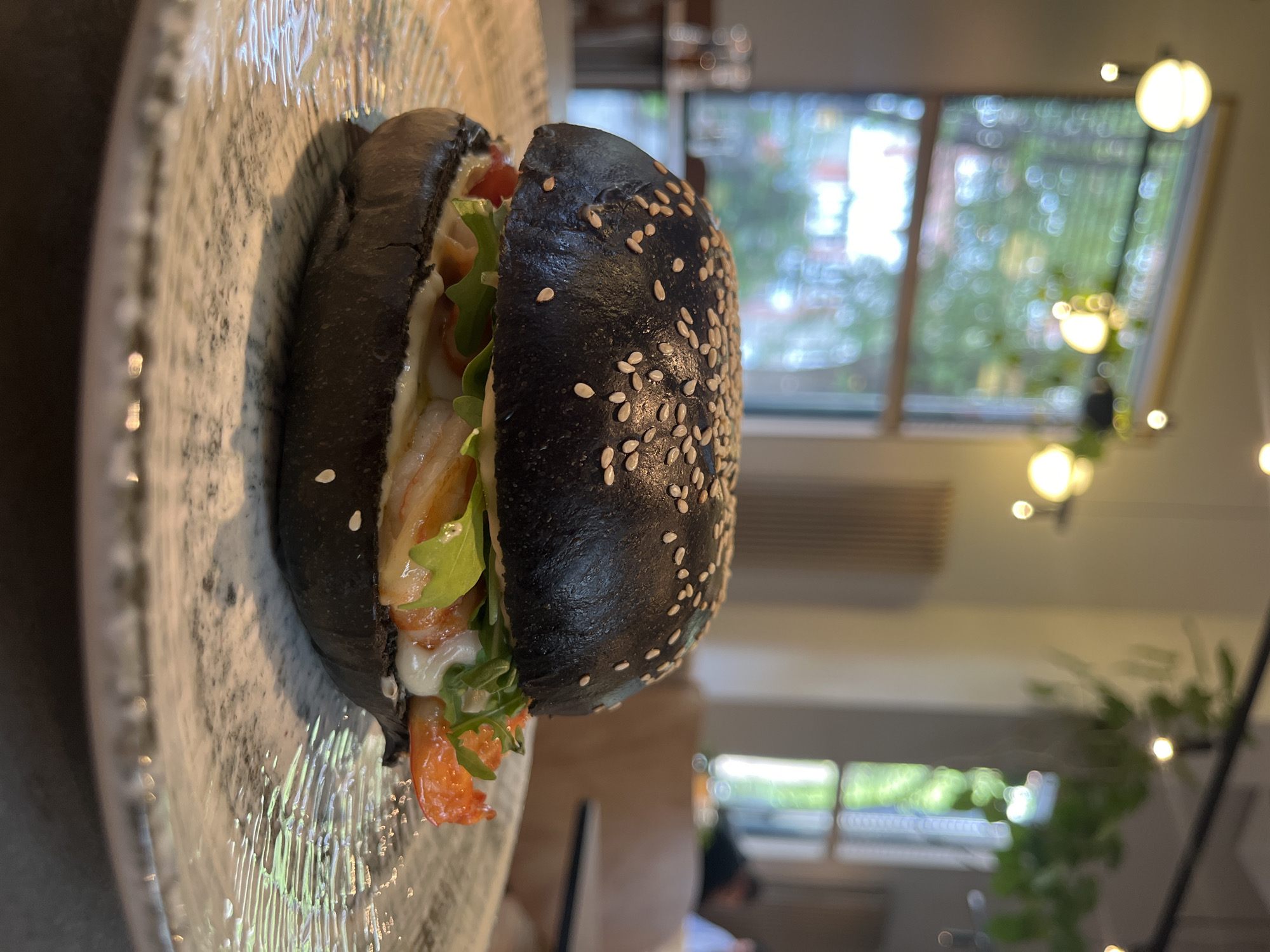 A close-up, rotated image shows a black sesame seed burger on a textured plate. The burger appears to contain lettuce, tomato, and what might be chicken or fish with a creamy sauce. The background includes a window with greenery visible outside and interior lighting fixtures that are out of focus. The image was likely taken indoors in an establishment or home in the city of Unknown, Unknown.