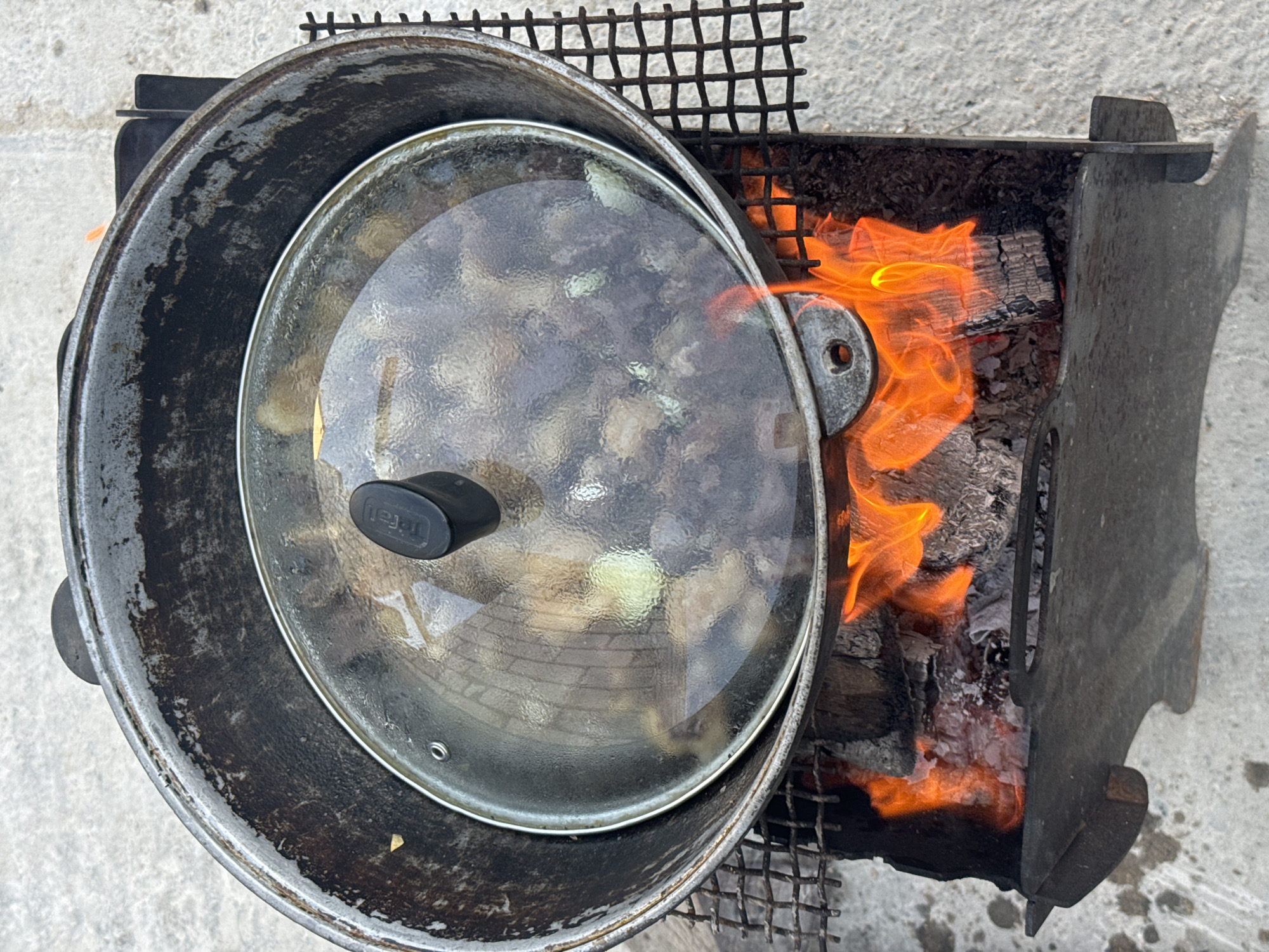 A black, used cast-iron pot with a clear glass lid is placed on a black metal grill over a wood fire. The pot is filled with food, which appears to be meat and vegetables. The fire is burning brightly with orange and yellow flames and embers. The grill is positioned over a black metal brazier containing the burning wood. The scene is set on a light gray, concrete surface. The background is out of focus and appears to be an outdoor setting. This scene is located in Atameken, Kazakhstan.