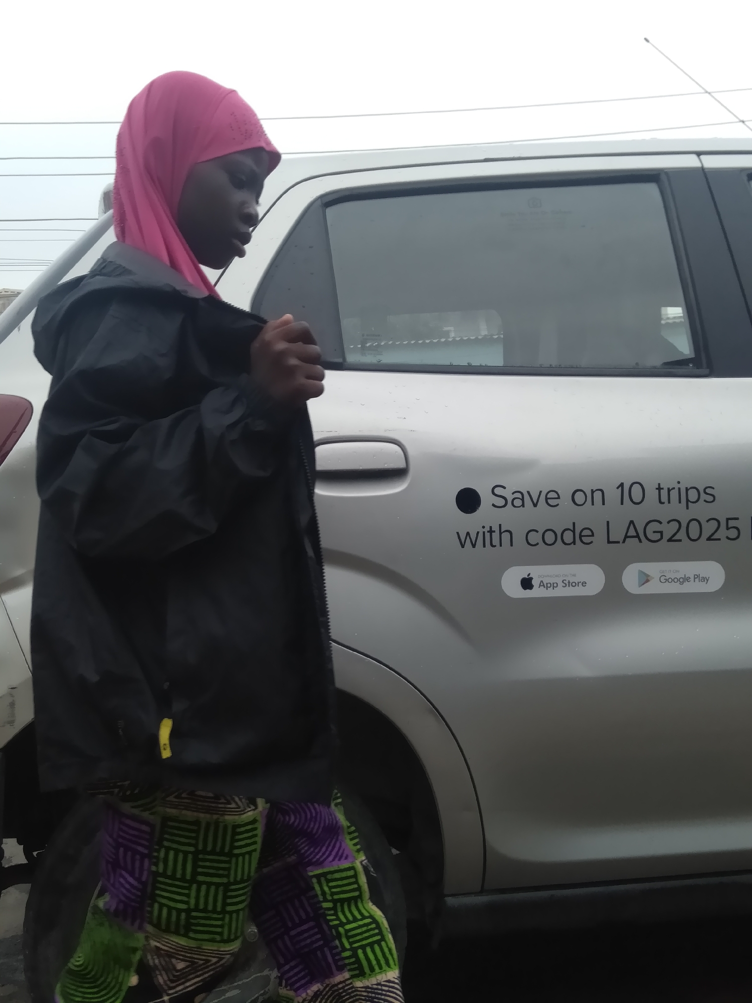 A photograph shows a person in the foreground wearing a pink headscarf, a black jacket, and colorful patterned pants standing next to a silver car. The car advertises "Save on 10 trips with code LAG2025" and displays logos for the Apple App Store and Google Play. The image has a slightly overcast or grey quality to it. The location is Shangisha, Nigeria.