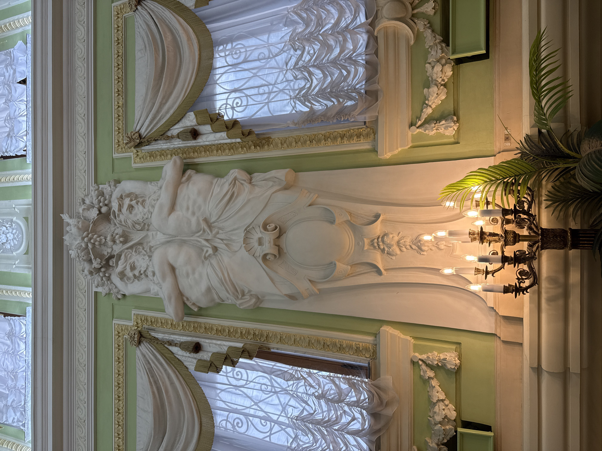 This is an interior shot of a room with ornate classical architecture, located in the Nizhny Novgorod Kremlin, Russia. The central focus is a large, white bas-relief sculpture depicting two muscular male figures intertwined, with a cluster of fruit and grapes above them. To the left and right of the sculpture are tall windows draped with heavy, cream-colored fabric that is gathered and tasseled. The window panes are covered with decorative metal grilles. The walls are painted a pale green, and elaborate white molding runs along the top of the walls and around the windows. In the foreground, a dark metal candelabra with seven lit electric candles stands on a tiered, cream-colored platform. A large, leafy green plant is positioned to the right of the candelabra, partially obscuring the lower section of the wall and the platform. The lighting in the scene appears to be artificial, with warm light emanating from the candelabra and illuminating the sculpture.