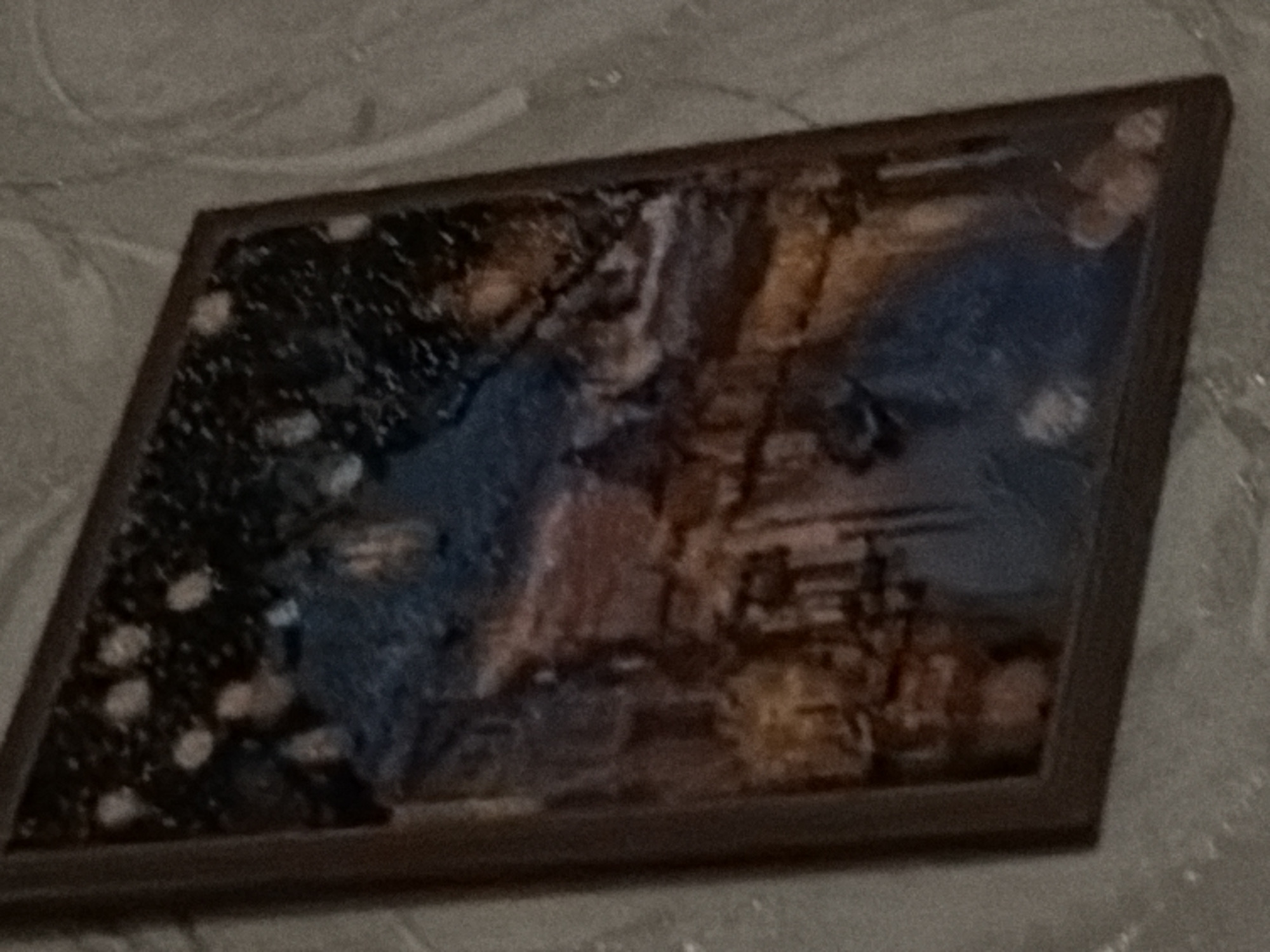 The image is a tilted, low-resolution photograph of a framed picture hanging on a textured wall. The picture itself appears to be a painting or a print of a nocturnal cityscape with a body of water in the foreground. The cityscape depicts buildings with illuminated windows and lights, possibly streetlights, reflecting on the water. The sky is dark with scattered points of light, suggesting stars or more distant lights. The colors are muted, with blues, oranges, and browns dominating. The wall has a faint pattern, possibly wallpaper. There is no indication of the city of Shakhty, Russia, or any other specific location within the image.