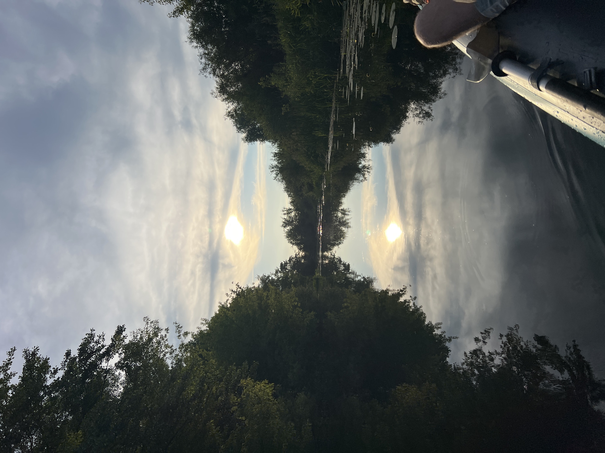 A view from a kayak on a calm river at sunset. The sun, low in the sky, reflects brightly on the water, creating a shimmering golden path. Both banks of the river are densely lined with lush green trees and reeds. The sky above is a blend of soft blues and grays, with streaks of white clouds catching the sunlight. In the foreground, part of the kayak and a person's legs are visible, indicating a passenger is present. The water is still, mirroring the trees and the sky with remarkable clarity.