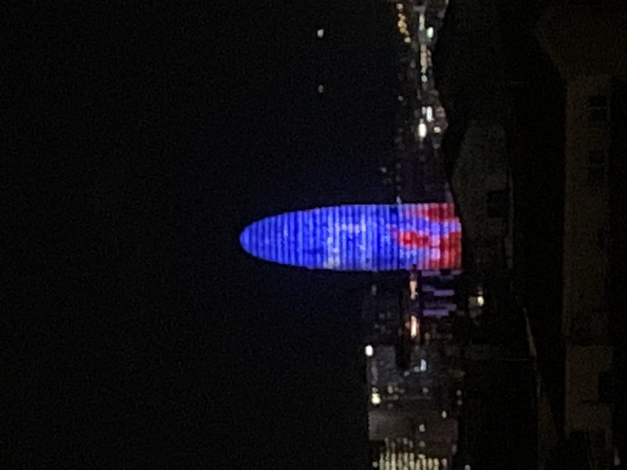 The image shows a night view of the Torre Glòries, located in Barcelona, Spain. The tower is illuminated with blue and red lights. Other city buildings are visible.