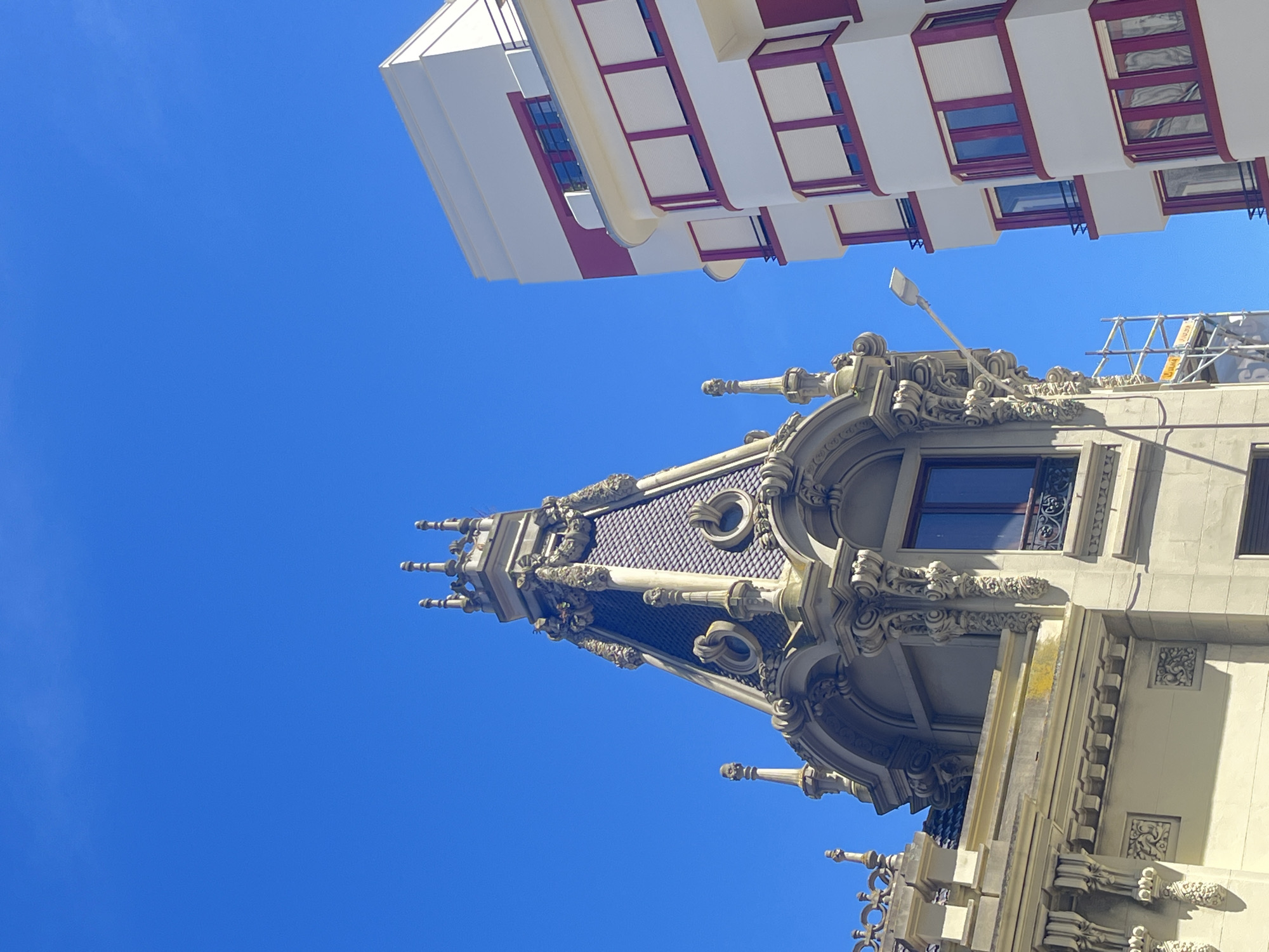 The image depicts a low-angle view of two buildings against a clear blue sky. One building is white with red window frames, and is positioned at the top of the frame. Below it is a light-colored building with elaborate architectural details, including turrets, ornate carvings, and a patterned roof. The buildings appear to be in A Coruña, Spain, based on the architectural style. There are scaffolding, light pole and ornate ornamentation visible.