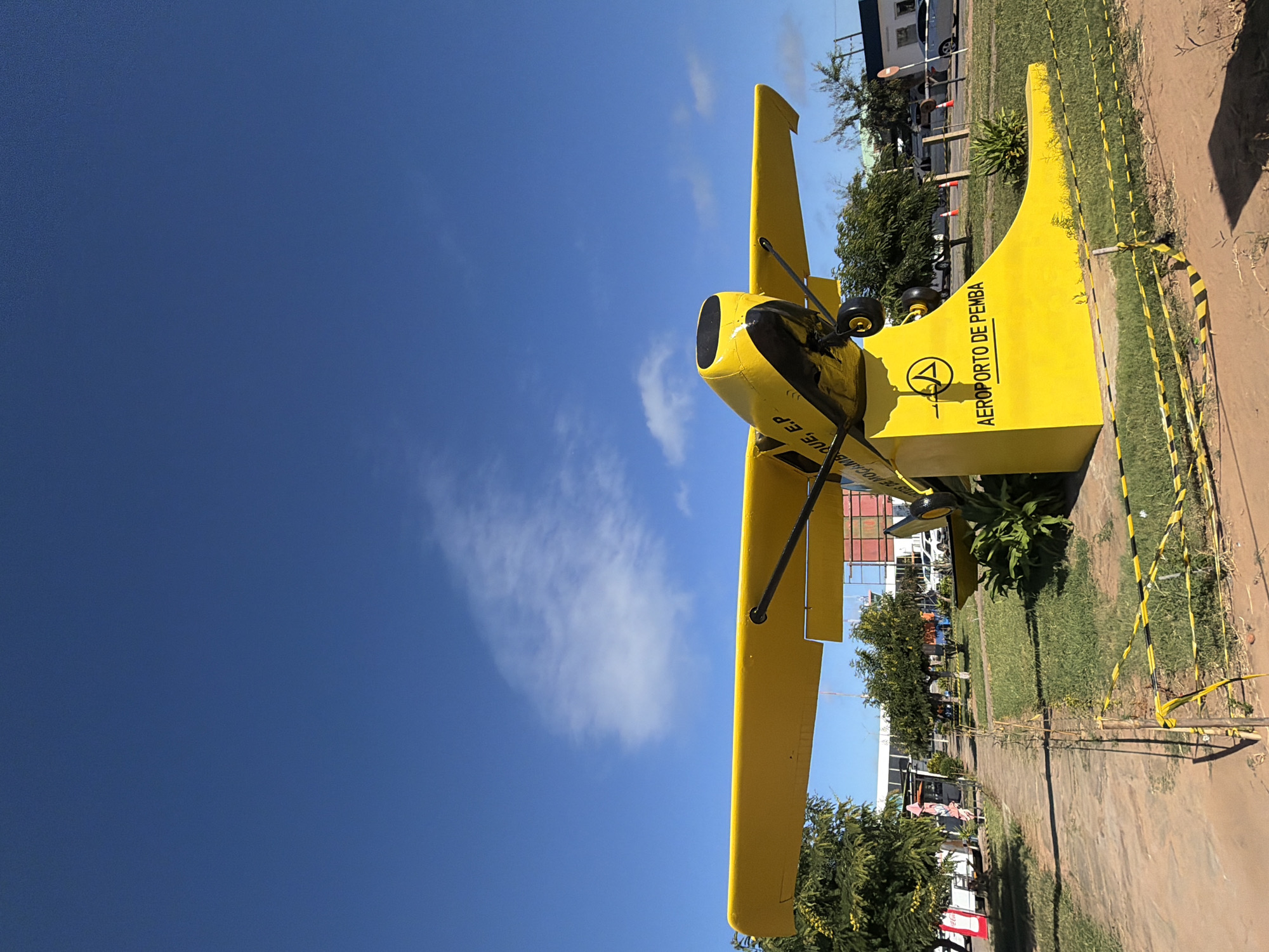 A large, bright yellow biplane is prominently displayed outdoors under a clear blue sky with a few wispy white clouds. The aircraft features black accents around its engine cowling and on its landing gear, contrasting with the vibrant yellow body and wings. Visible struts connect the biplane's wings.
The plane serves as a static monument or landmark, placed on a patch of green grass beside a dirt path. Around the base of the display, black and yellow striped barrier tape is present, suggesting a restricted or designated area. In the background, there are several green trees and bushes, along with buildings that appear to be part of an airport terminal or facility. Parked vehicles, including cars and vans, are visible near the buildings. A few indistinct human figures can be seen in the distance, walking or standing, but they are not central to the image.
The time of day is daytime, evidenced by the bright sunlight and clear visibility, casting some shadows.
Visible text on the side of the biplane's tail section clearly reads "AEROPORTO DE PEMBA," indicating Pemba Airport. Another visible fragment of text below the cockpit area reads "MOCAMBIQUE E.P," identifying the location as Mozambique. A circular logo containing an "A" is also present next to the airport name.