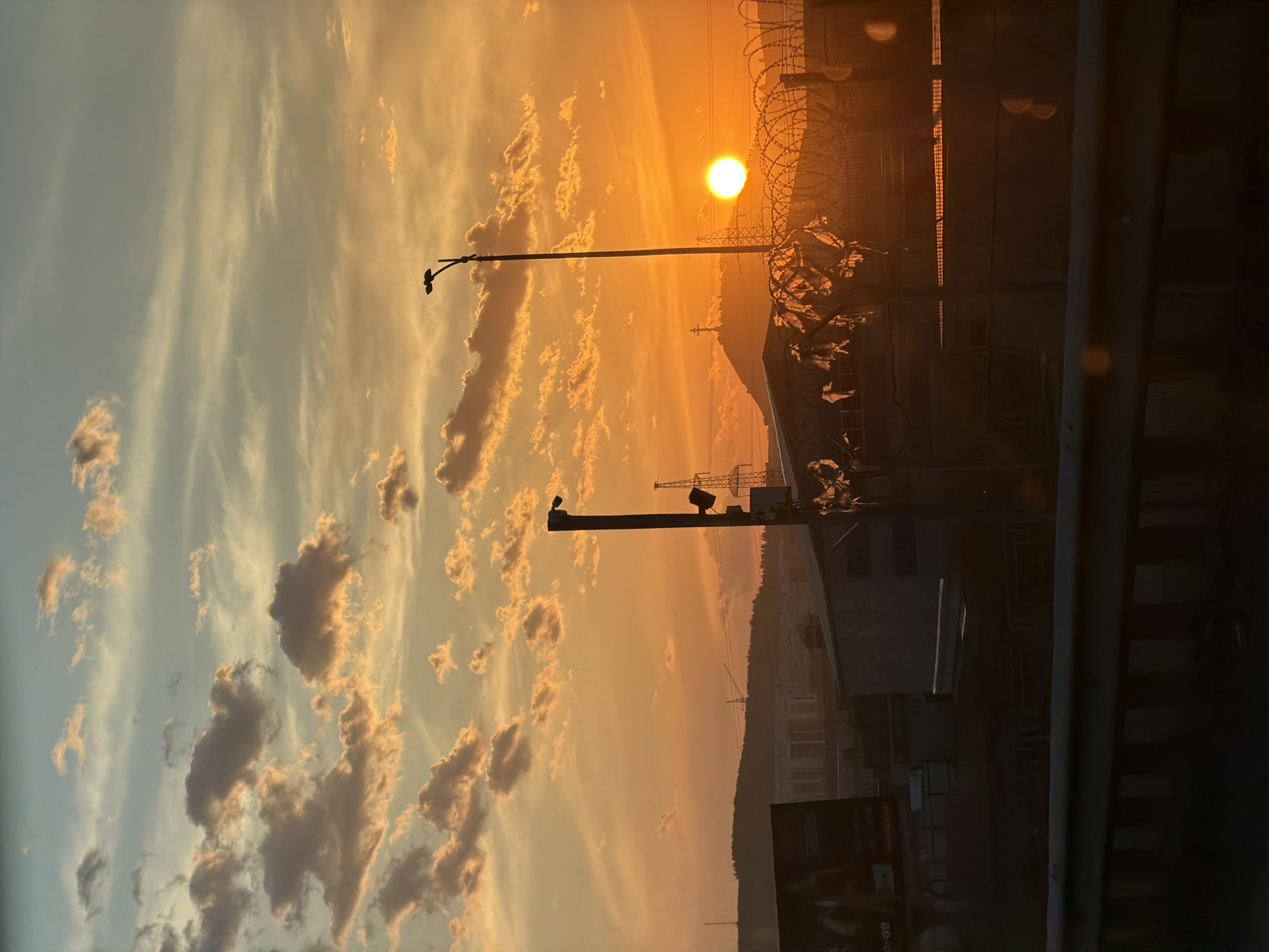 The image depicts a sunset scene over Krasnoyarsk, Russia. The sun is visible, positioned near a fence topped with barbed wire. A street light stands in the foreground. The sky is filled with clouds, tinted orange and yellow by the setting sun. Some buildings can be seen in the background, though the details are obscured. The scene is viewed from a slightly obscured vantage point, potentially from inside a building or vehicle, indicated by the dark areas on the edges of the frame.
