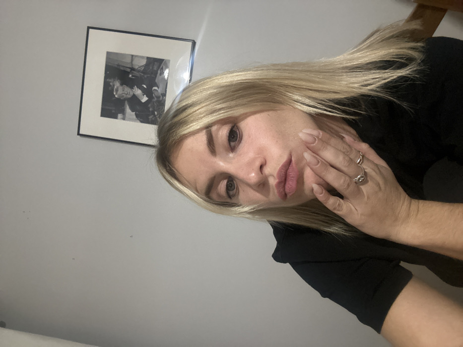 A woman with blonde hair and light skin is shown in a close-up. She wears a black shirt and multiple rings on her fingers. Her hand is near her cheek. The background is a plain, light gray wall. A framed, black and white picture hangs on the wall behind her. The photo appears to depict men in suits. The image is slightly tilted.