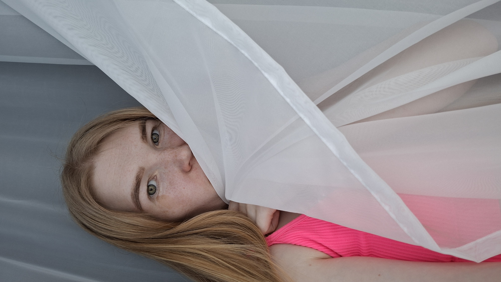 A fair-skinned woman with freckles and light brown hair is lying on a gray surface, possibly a bed or floor, in an indoor setting. She is partially obscured by a translucent white fabric, draped over part of her face and body. The woman is wearing a bright pink top. The lower portion of her face, including her nose and mouth, are visible below the fabric, while her eyes are visible above. Her hair is spread out on the gray surface. There are no distinguishing characteristics that indicate the location as Chaikovsky, Russia.