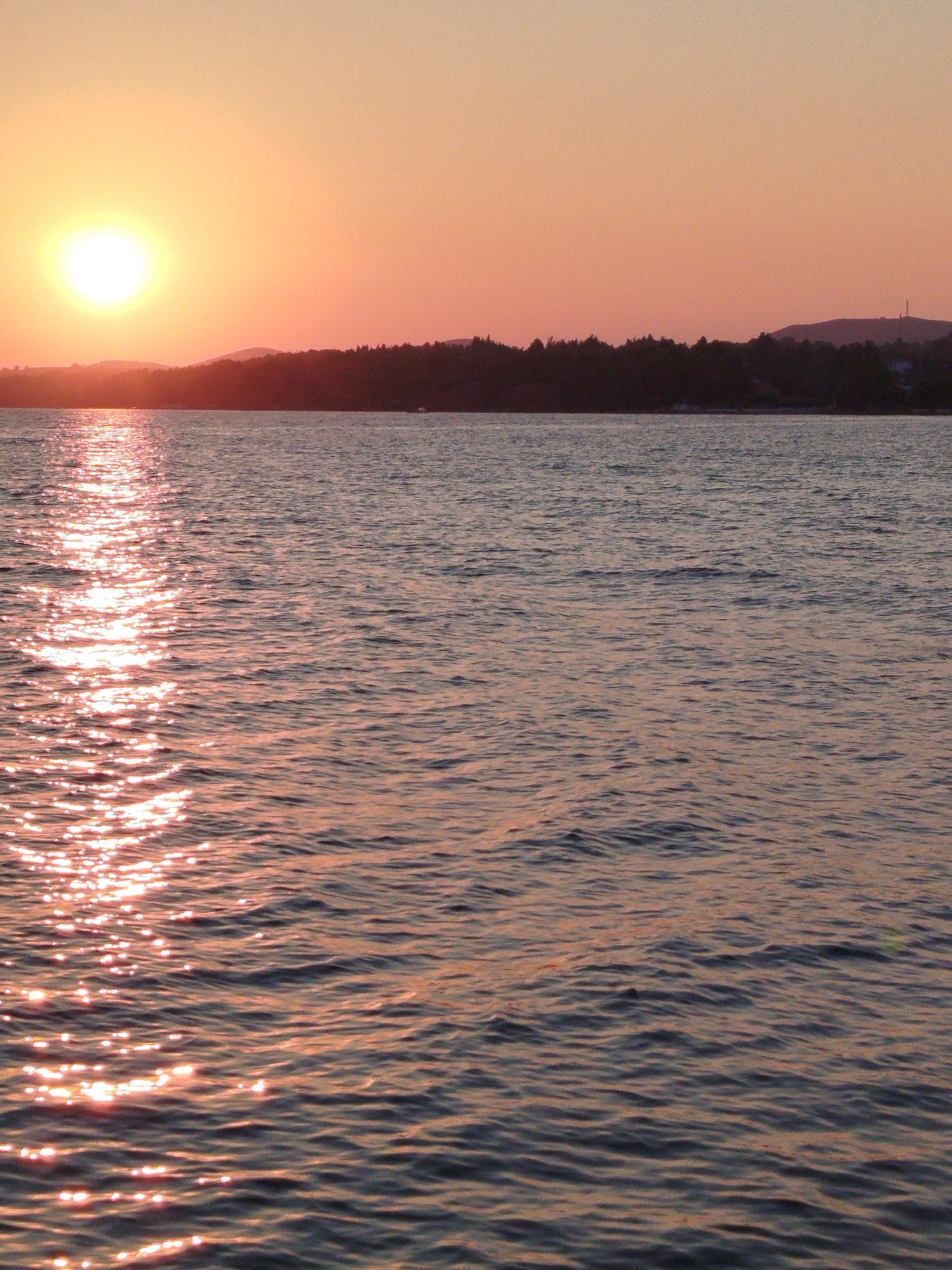 A vertical, close-up shot captures the sea at Sithonia Municipal Unit, Greece, during sunset. The sun, a bright white orb, is visible in the upper left quadrant, casting a shimmering path of light across the rippling water. The horizon is lined with a dark silhouette of trees and hills, with a faint tower visible on the rightmost hill. The sky is a gradient of pale orange to pink, reflecting the warm hues of the setting sun. The water is a deep blue, with ripples and small waves disturbing its surface, catching the sunlight in glittering highlights.