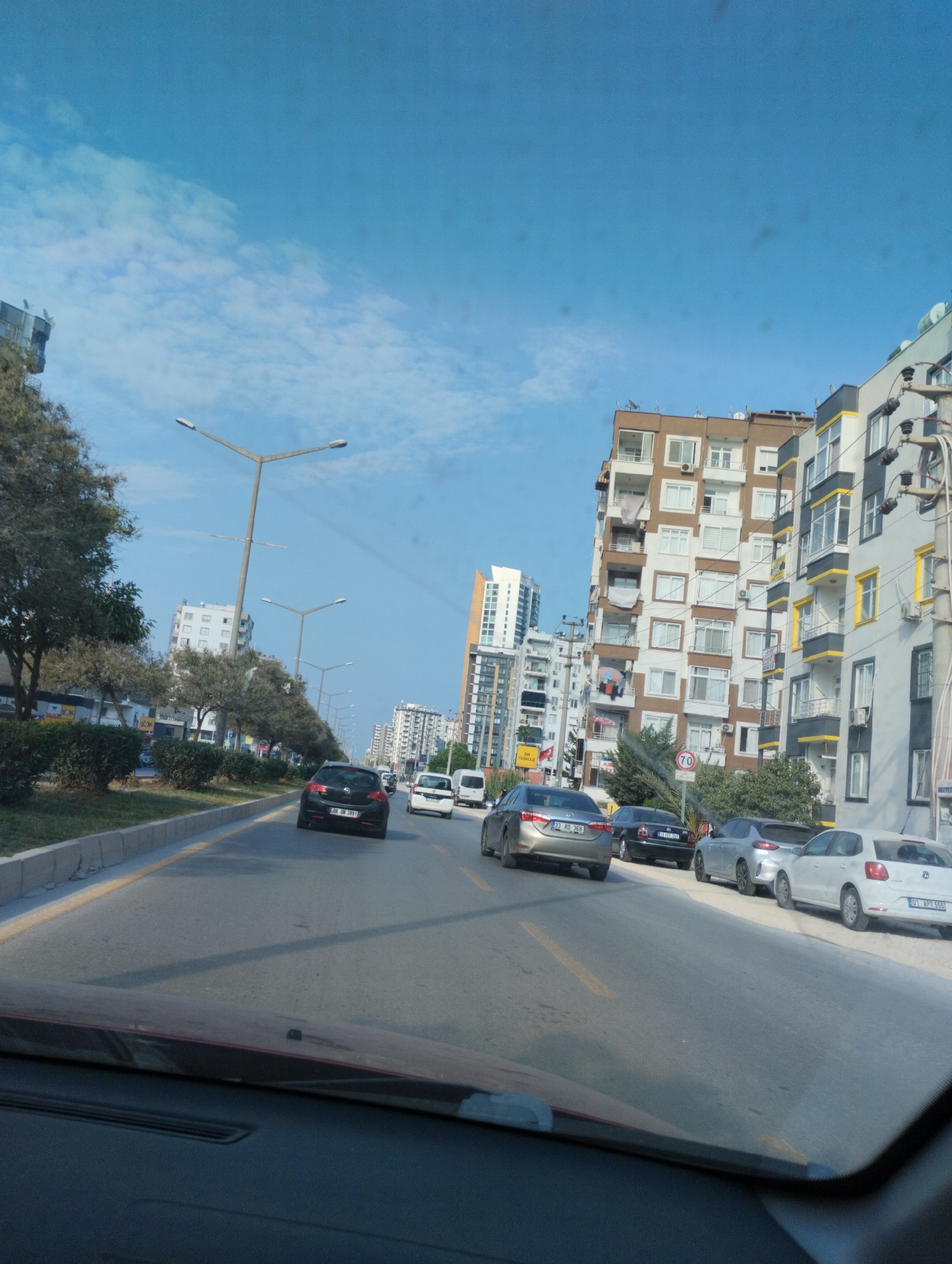 The image depicts a street scene in Mezitli, Turkey, viewed from the interior of a vehicle. The road is paved with asphalt and divided by yellow lines. Several cars are visible on the road, including a black hatchback, a silver sedan, and a white hatchback. The cars are in motion or parked. Along the right side of the road, there is a sidewalk and a row of parked cars. The left side of the road features a median with trees and bushes. In the background, a series of apartment buildings of varying heights and architectural styles line the street, suggesting an urban environment. The sky is blue with scattered white clouds. Streetlights are also visible along the road. The overall scene conveys a typical day in a city with moderate traffic and residential buildings.