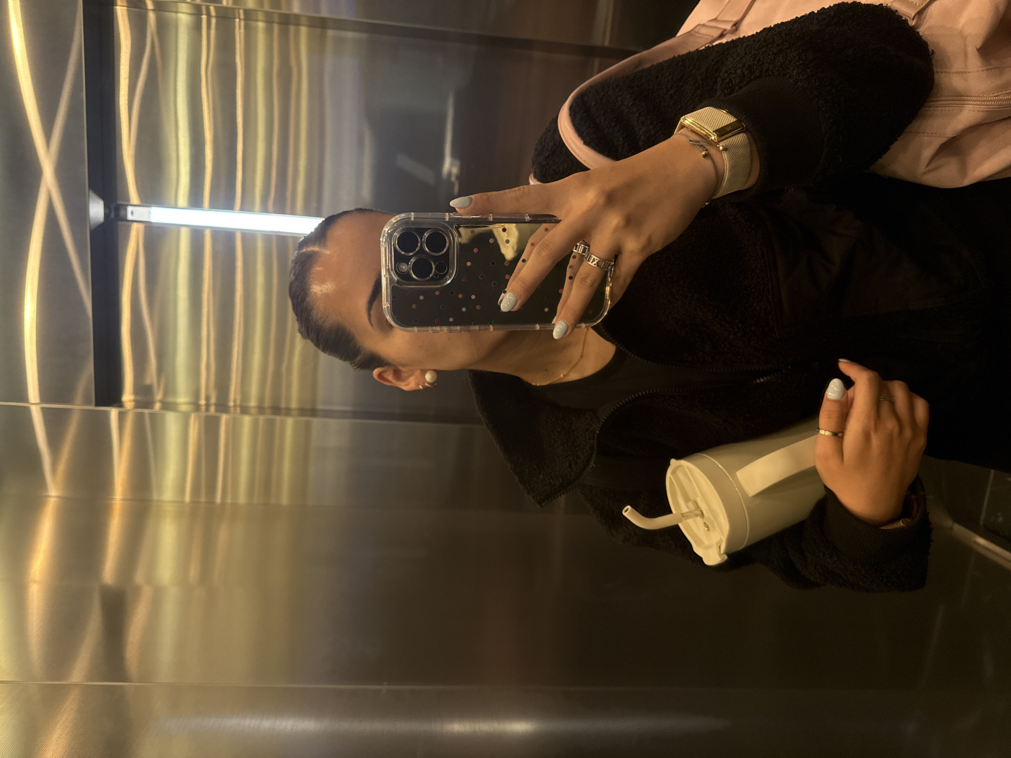 A person is captured in a mirror selfie, reflecting off the highly polished, metallic walls of what appears to be an elevator or a modern indoor space. The reflection shows the individual's right side, with their hair pulled back and a small white stud earring visible.
Their right hand is prominently holding a smartphone, likely an iPhone, encased in a clear cover adorned with small pink and black polka dots. The person's fingernails are painted a light blue. On their fingers, several rings are worn: a plain band on the ring finger, and more decorative rings on the middle and index fingers, one featuring what looks like Roman numerals. A light-colored watch with a rectangular, gold-toned face and a woven or mesh band is on their wrist, along with a delicate bracelet.
The individual is wearing a black, textured jacket, possibly fleece or sherpa, and a light pink bag or garment is visible over their left shoulder. In their left hand, they hold a light-colored, possibly pale blue or white, tumbler or water bottle with a straw. The metallic walls of the setting reflect a horizontal strip light, creating bright, elongated reflections across the surfaces. The scene is brightly lit by artificial light. While the location context of Puebla, Mexico is provided, there are no specific cues within the image itself to confirm this. No discernible text is visible on any objects or surfaces.