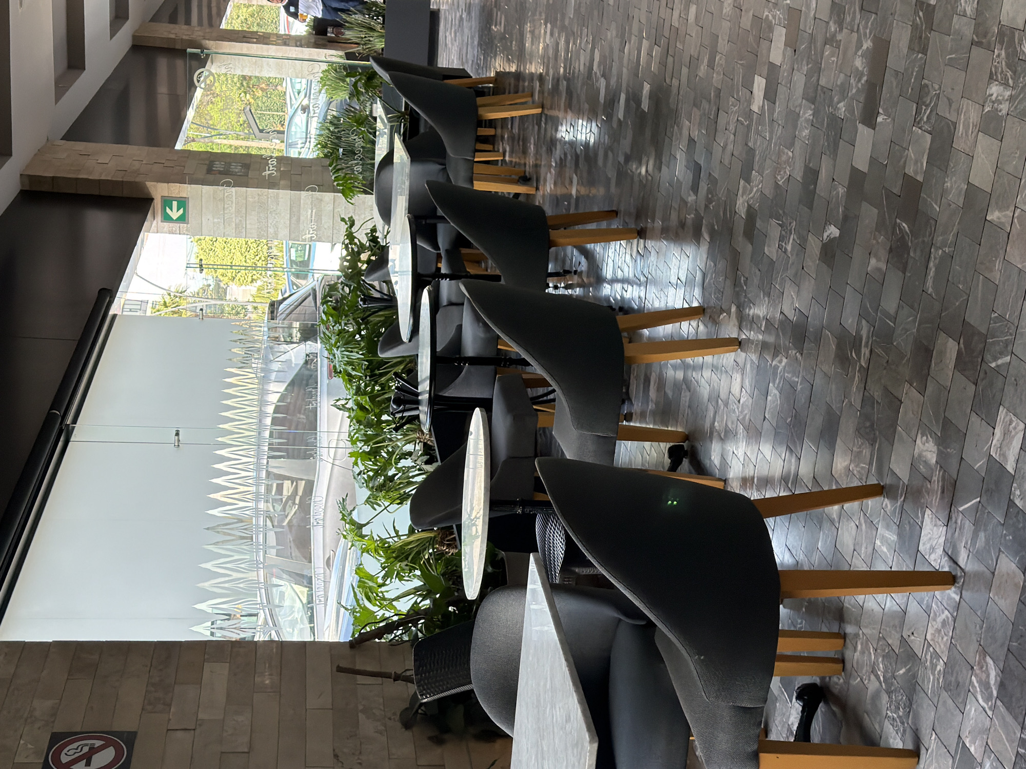 The image displays a modern indoor seating area, likely a cafe or restaurant, illuminated by natural light. The space is arranged alongside a large glass partition that offers a view of an outdoor street scene. The seating comprises numerous dark gray upholstered chairs with light-colored wooden legs, positioned around various light-toned, either round or rectangular, tables. A continuous row of vibrant green potted plants serves as a natural divider between the seating and the expansive glass wall.
The floor features a distinctive pattern of dark gray rectangular tiles laid diagonally. An interior wall is clad with light brown, textured stone tiles, adding an earthy element to the design. Through the clear upper section of the glass partition, a bright daytime setting is visible, showing lush green trees, parked cars, and a partially obscured individual in the distance. The lower half of the glass partition is frosted with an opaque, decorative zig-zag pattern, resembling a mountain range.
Visible text includes a green down arrow displayed on an interior column and a circular "NO SMOKING" sign, which also clearly states "NO FUMAR" in Spanish, located on the stone-clad wall in the foreground. Reversed reflections on the glass partition show words such as "CHOCOLAT" and other stylized script, possibly indicating nearby establishments or decorative elements. The chairs appear empty, suggesting the area is not currently busy. The overall impression is a contemporary, well-lit space in Puebla, Mexico, characterized by its clean aesthetics and connection to the outdoors.