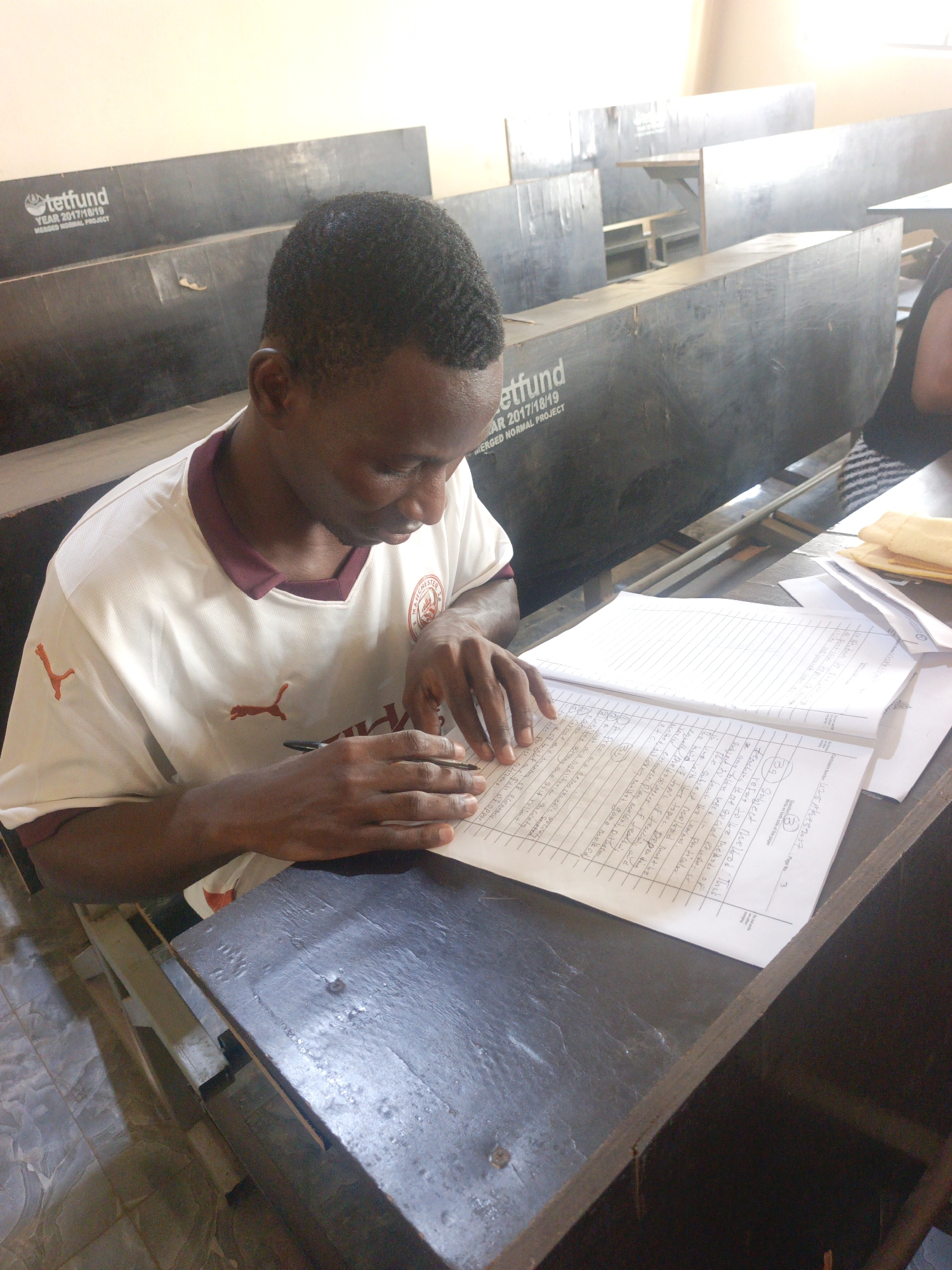 A young man is seated at a desk in a classroom, writing in a notebook. He is wearing a white t-shirt with a Puma logo and a maroon collar. The notebook is open and filled with handwriting. On the desk, there are also some loose papers. The desk is dark and worn, and the classroom has rows of desks behind him, some with "tetfund YEAR 2017/18/19 MERGED NORMAL PROJECT" printed on them. The lighting is bright and possibly natural, coming from the upper left of the frame.