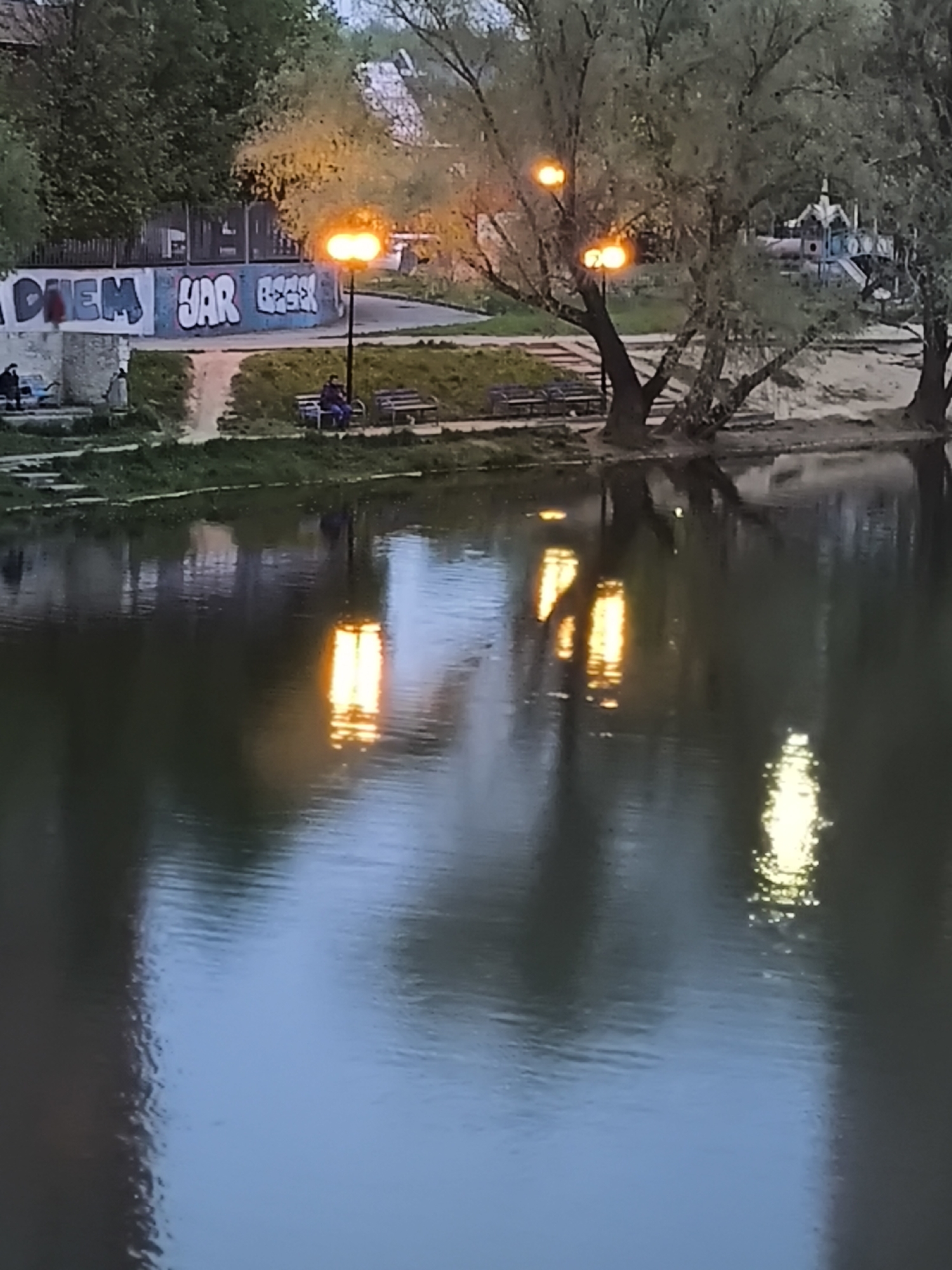 The image shows a pond at dusk in Khimki, Russia. The still water reflects the orange glow of streetlights positioned along the bank. The bank features a concrete wall with graffiti, trees, and benches where people are seated. A playground structure is visible in the background.