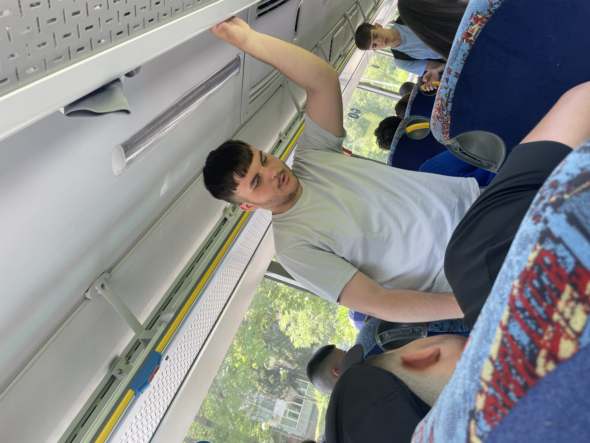 The image shows the interior of a bus, captured from a tilted, upward-looking perspective. The scene appears to be set during the daytime, with bright natural light illuminating the cabin and verdant green trees visible through the windows, suggesting a sunny day.
The primary subject is a young man standing in the aisle, wearing a plain light grey t-shirt. He has short dark hair with a blunt fringe and a light beard and mustache. His left arm is extended upwards, grasping an overhead grab bar for support. His facial expression is neutral.
Several other passengers are visible. In the background, another young man with short hair, wearing a light blue t-shirt and a black backpack, is seated and looking towards the front of the bus. Closer to the foreground, the back of a dark-haired person's head is visible, and partially obscuring the lower right, the ear and back of the head of a person wearing a black cap can be seen.
The bus interior features typical elements: overhead perforated panels, a shiny silver grab rail, and blue bus seats with a distinct pattern of red, yellow, and blue geometric shapes. A yellow safety bar runs along the window line, fitted with a small blue component. The windows reveal a clear view of the outside, dominated by foliage. No discernible text is visible on any objects or surfaces within the bus. The image depicts a routine journey on public transport, likely within Burgas, Bulgaria.