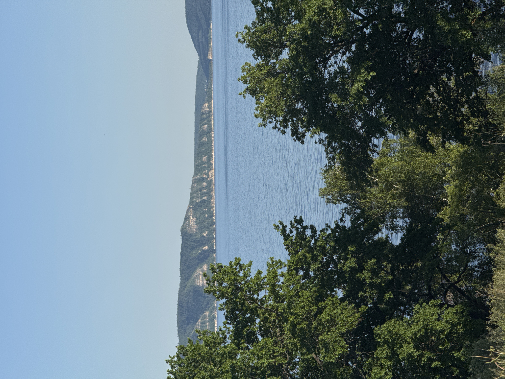 The image depicts a body of water with a land formation on the left side and trees on the right. The sky is a light blue. The location and city are unknown.