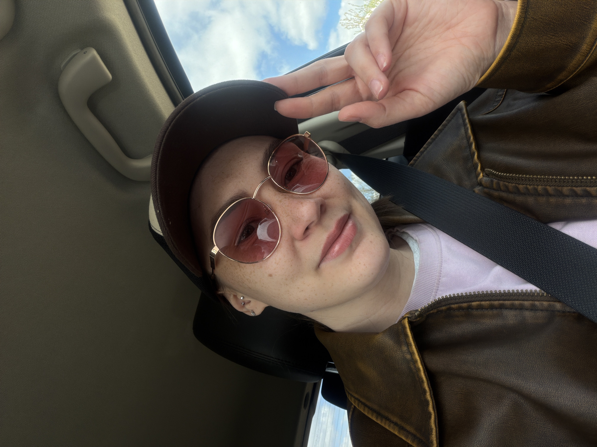 A woman is sitting in the driver's seat of a car, wearing a brown leather jacket, a light purple shirt, and a brown baseball cap. She also wears pink-tinted sunglasses. She is making a peace sign with her right hand. A black seatbelt is fastened across her chest. The interior of the car is visible, including the car's roof, side panel, and a handle on the ceiling. The background shows a sky with clouds. It is not possible to determine the specific location in Chaikovsky, Russia from the image.