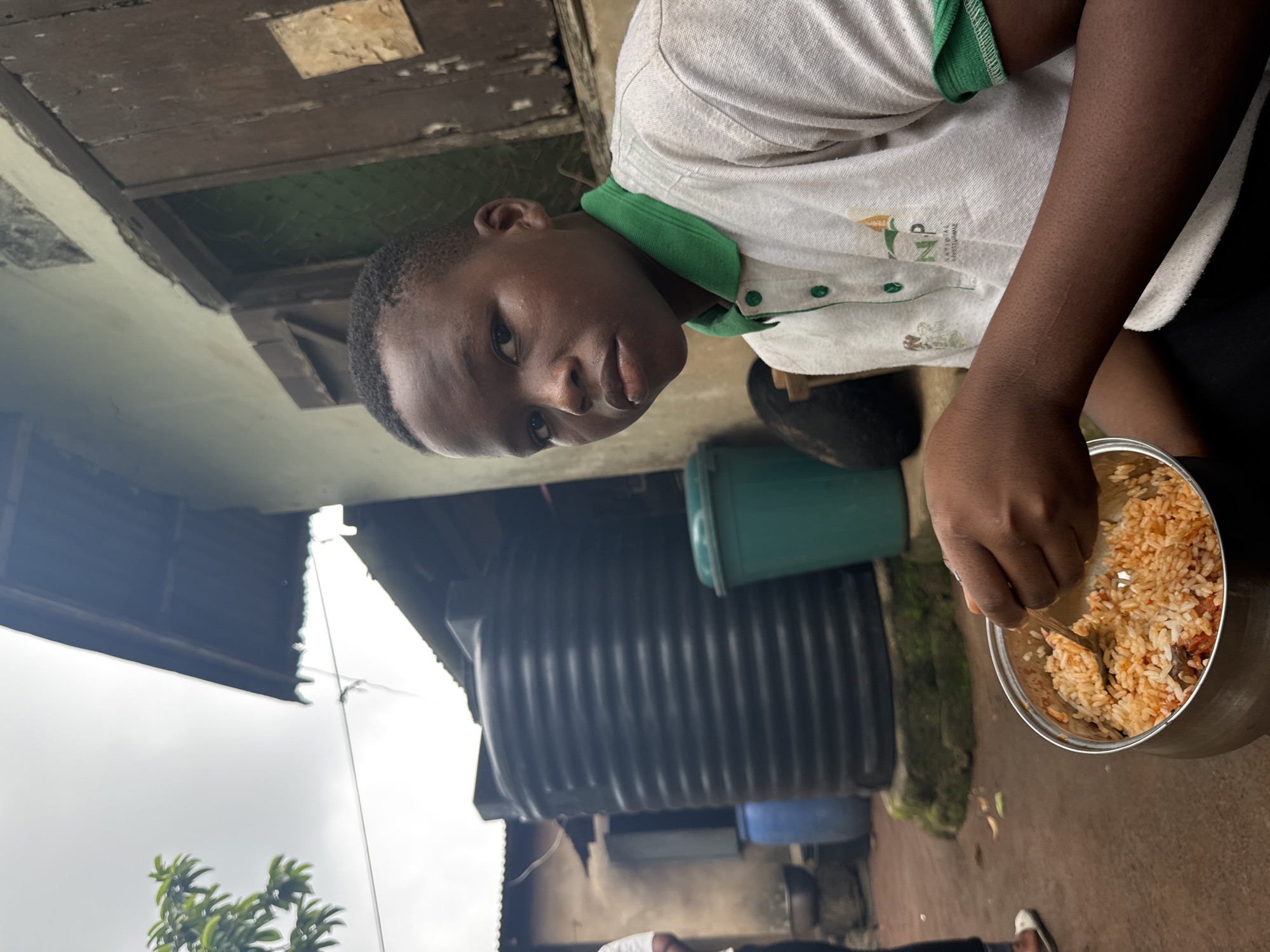A young person is shown outdoors, holding a pot of rice and stirring it with a spoon. The individual has dark skin, short hair, and wears a white short-sleeved shirt with a green collar. A dark water tank is behind the person. The environment includes a wooden structure, plants, and a cloudy sky is visible in the background. The image suggests the setting is Aboh, Nigeria, inferred through contextual clues such as the water tank and architecture.