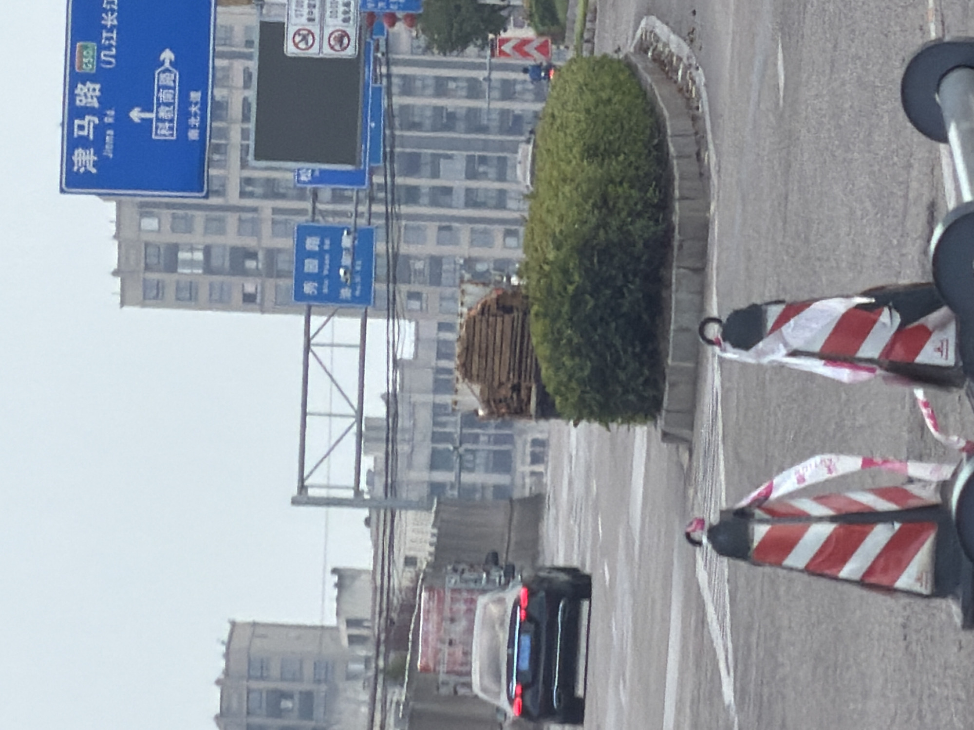The image captures a street scene in Jiangjin, China, under an overcast sky. In the foreground, two traffic cones, striped red and white with reflective tape, stand on the asphalt road, one with a white plastic ribbon draped over it. Beyond them, a black sedan with illuminated red taillights is positioned on the road, facing away from the camera. To the right of the sedan, a large pile of lumber is stacked on a flatbed trailer or truck.
Further back, the environment is characterized by various buildings and infrastructure. A large, curved hedge is situated in the midground, bordered by a concrete retaining wall. Overhead, a blue highway sign with Chinese characters and the English translation "Jioma Rd." is visible, indicating directions to "Southeastern South Road" and "North-South Avenue." Another blue sign below it reads "Xiangyun Road." In the background, multi-story residential buildings with numerous windows dominate the skyline. Traffic lights and other street signs are also present. The overall impression is that of an urban environment undergoing some form of road construction or modification, given the presence of the lumber and traffic cones.