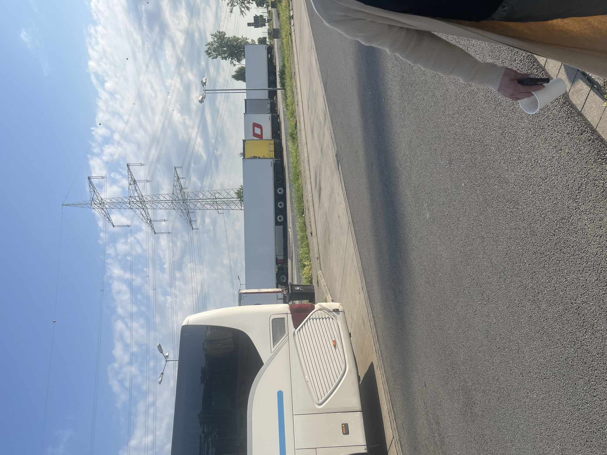 In Eschweiler, Germany, on a clear day with a blue sky dotted with white clouds, a white bus is parked on the left side of the frame. To the right of the bus, a line of semi-trailer trucks is parked. One truck has a yellow cargo box, and another has a red logo on its white cargo box. A person, partially visible on the right side of the image, is holding a white cup and a dark object, possibly a phone, in their hand. In the background, tall electrical pylons and streetlights are visible, along with trees. The scene appears to be a roadside rest stop or parking area.