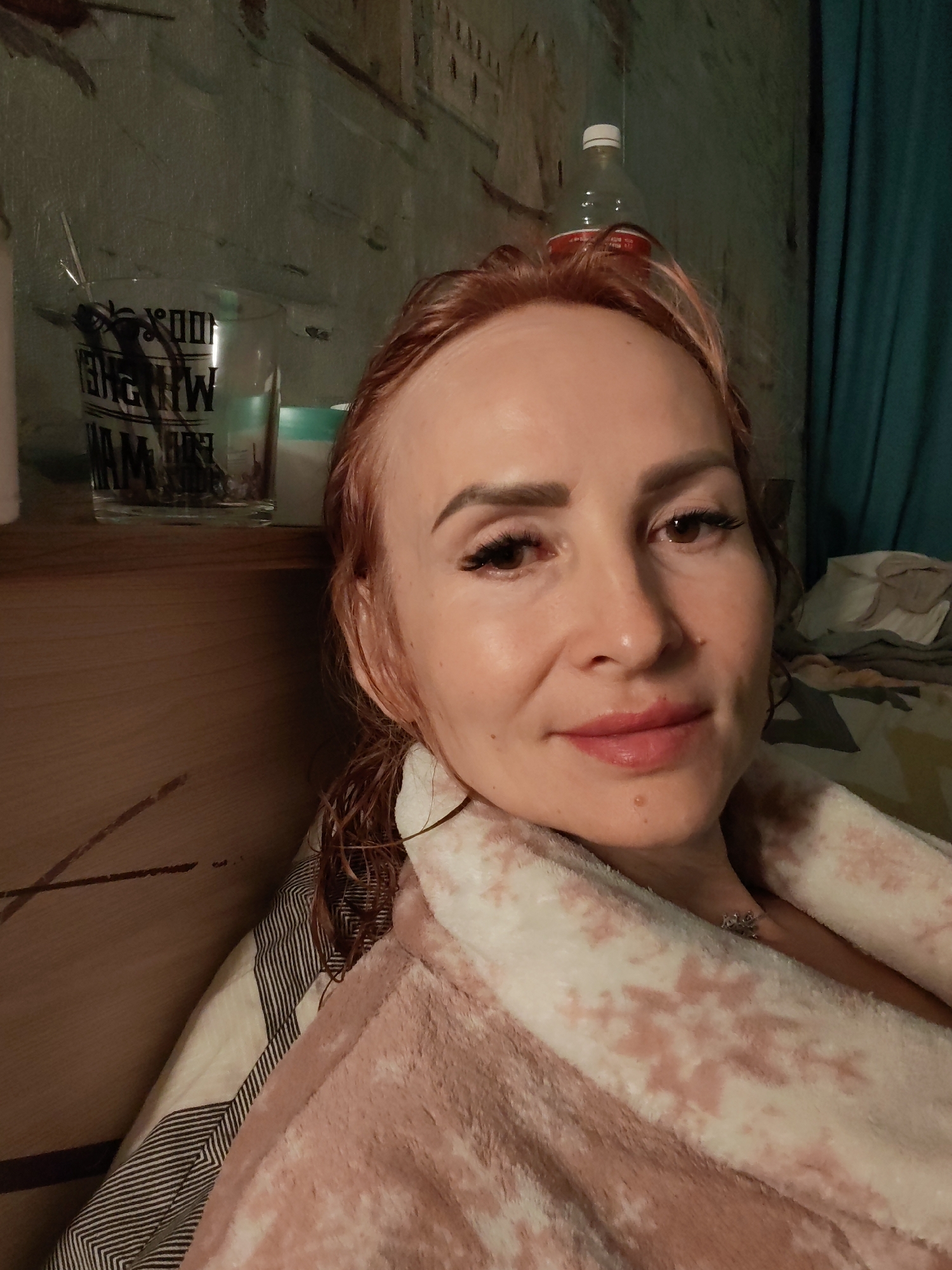 A medium shot captured from a slightly high angle shows a woman with light skin and reddish-brown hair, appearing to be in her late 30s or early 40s, looking towards the camera. She is wearing a light pink bathrobe with a snowflake pattern. Her hair is damp and pulled back, with some strands falling forward. She has full lips with a touch of pink lipstick and her eyebrows are well-defined. Her right eye is slightly closed, and she is wearing false eyelashes. A small mole is visible on her chin.
The background suggests an indoor setting, likely a bedroom or living area. To the left, a wooden shelf holds a clear glass with text that appears to be in a foreign language and some dark objects inside, possibly a decorative spider. Behind the glass, there's a glimpse of a green and blue wall with a textured pattern that resembles a city skyline or building facades. To the right of the woman's head, a plastic bottle with a white cap and a red label sits on top of what seems to be the wall or a decorative element. Further to the right, a teal-colored curtain hangs. In the lower right portion of the image, a patterned blanket or duvet is visible, with a geometric black and white striped pattern and a light-colored fabric.
The lighting in the image is dim, casting a soft glow on the woman's face. There are no immediate indications that this scene is located in the city of Chaikovsky, Russia, or any specific city. The environment appears to be a personal living space.
