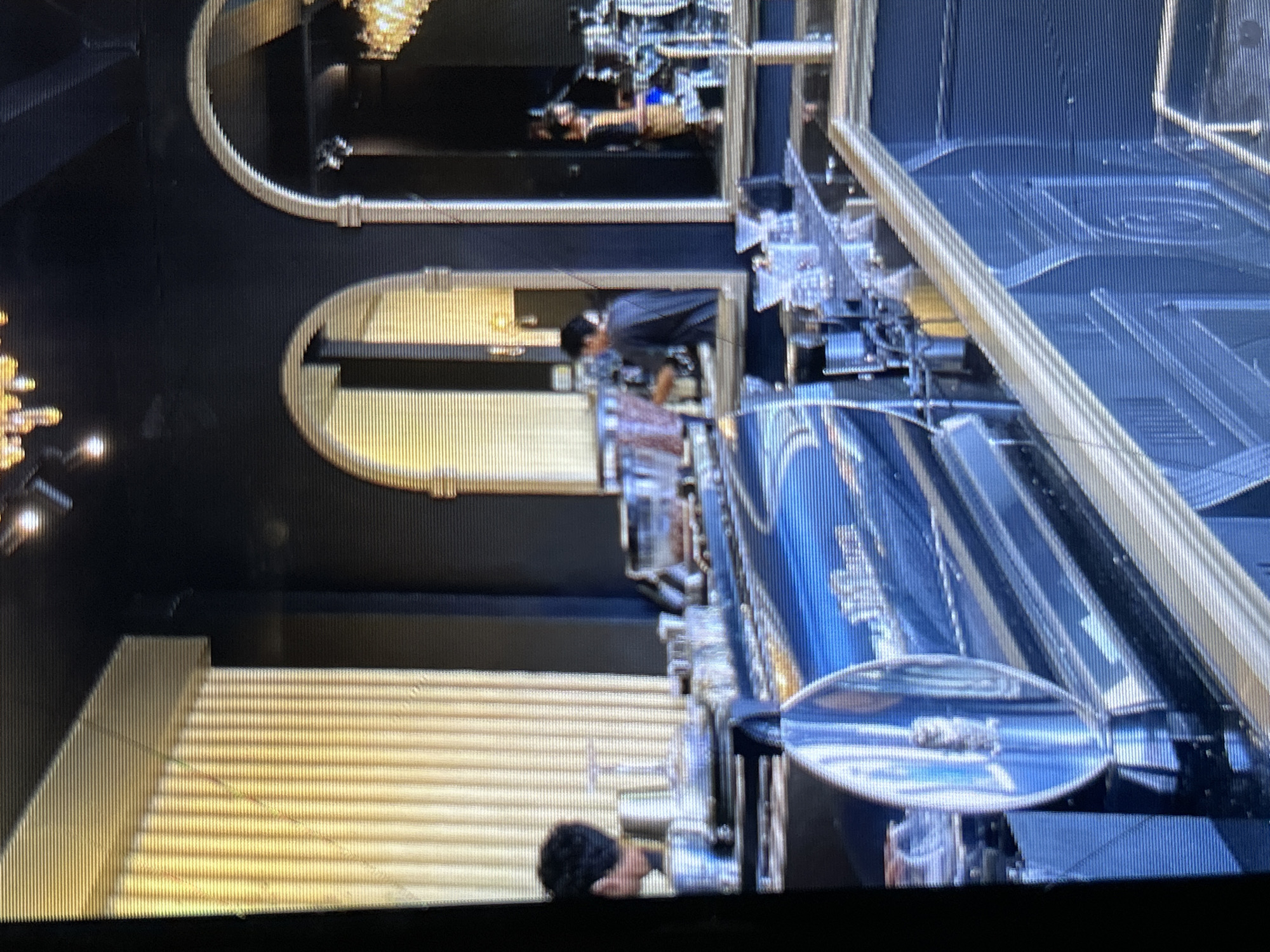 The image shows the interior of a cafe with a prominent bar area. The bar is made of a dark, reflective material with gold accents. Behind the bar, there are several baristas at work. One barista, with dark hair, is visible on the left side of the image, wearing a dark shirt. Another barista is in the background, also wearing a dark shirt. The cafe has an arched entrance on the right side, and chandeliers are visible on the ceiling. The walls appear to be dark, and there are vertical, golden textured panels on the left side. Various coffee-making equipment, cups, and containers are arranged on the bar. The lighting is dim and atmospheric, with some spotlights illuminating areas of the bar. The overall impression is of a modern and stylish cafe setting. The location is specified as Depok, Indonesia.