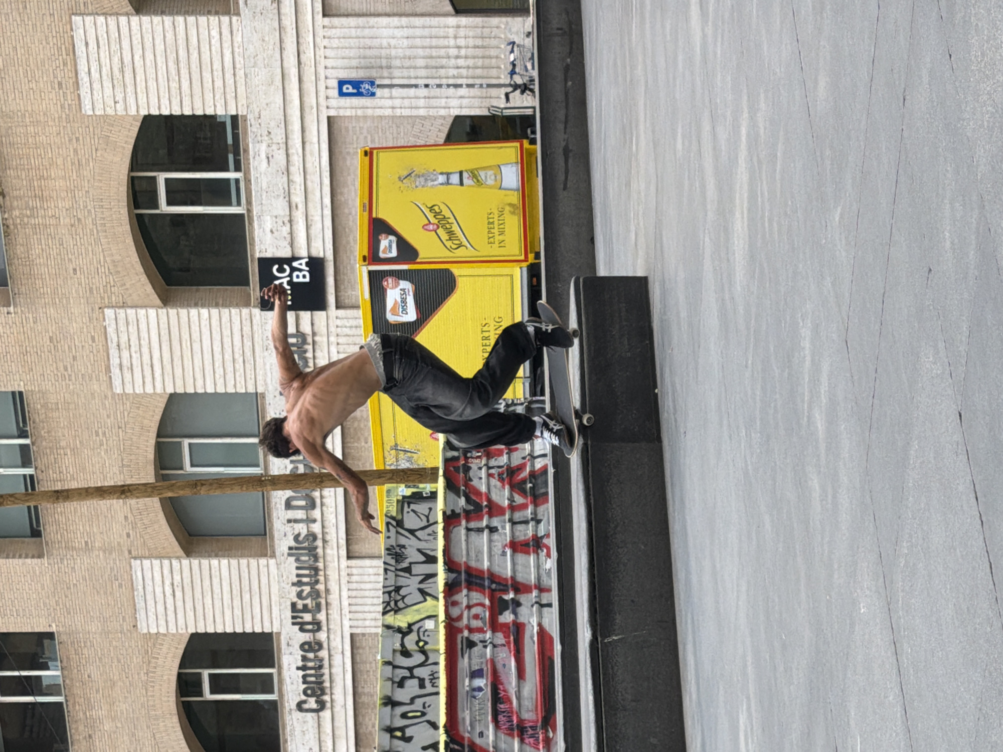 A dynamic urban scene captures a shirtless male skateboarder mid-trick, performing a grind or slide on a dark grey ledge. He is lean, with dark hair, wearing dark pants and sneakers, and extends his arms for balance. His skateboard is visible beneath his feet on the edge of the structure.
In the background, a large, multi-story building features light-colored, possibly brick or stone, facade with arched windows and vertical paneling. The windows are dark, reflecting little. In front of this building, to the right of the skateboarder, are two large yellow structures, likely advertising kiosks or containers. These feature prominent "Schweppes" branding with a bottle illustration and the text "EXPERTS - IN MIXING". Another logo, "DISBESA", is also visible. A blue street sign with a white 'P' and bicycle icon can be seen further back.
A thin, dark pole runs vertically through the middle ground. The side of the ledge facing away from the viewer is covered in red, black, and white graffiti. On the building facade, partial text "Centre d'Estudis i D..." and "AC BA" on a black sign are visible. The ground surface in the foreground and to the right is a light grey, textured material, possibly concrete or asphalt. The lighting suggests daytime, with soft, diffused light, indicating an overcast day or shaded area. The overall setting is an urban environment, consistent with Barcelona, Spain.