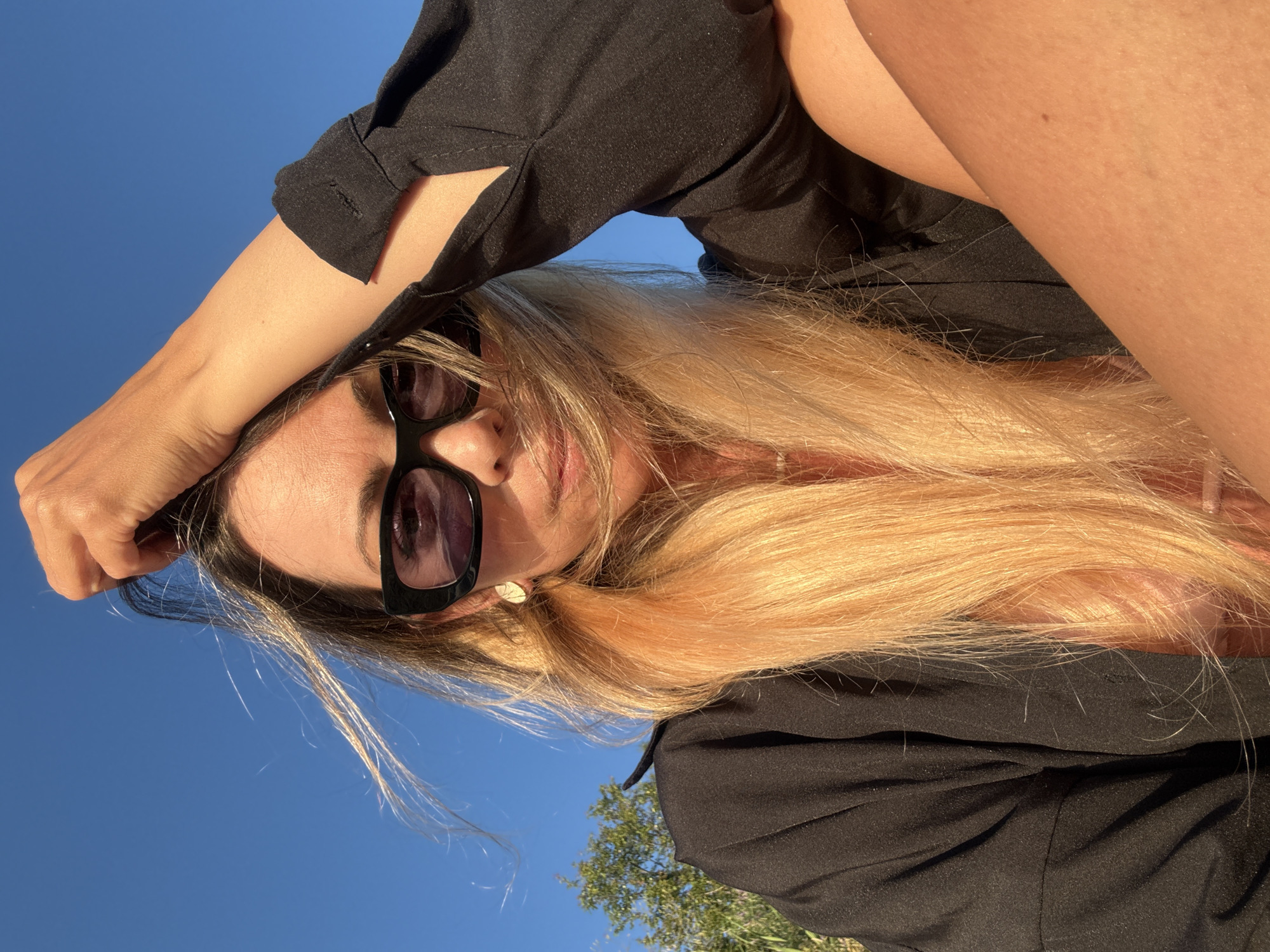 A woman with long blonde hair, wearing sunglasses and a black top, poses for a selfie outdoors. Her left arm is raised, touching her forehead, and her right arm is visible on her right side. The background is a clear blue sky, with a hint of green foliage at the bottom of the frame, suggesting an outdoor setting. The sunlight creates a warm glow on her skin and hair, indicating it is daytime, likely late afternoon or early morning due to the angle of the light. The overall mood appears relaxed and confident. The image appears to be taken in Nesebar, Bulgaria. There is no visible text in the image.