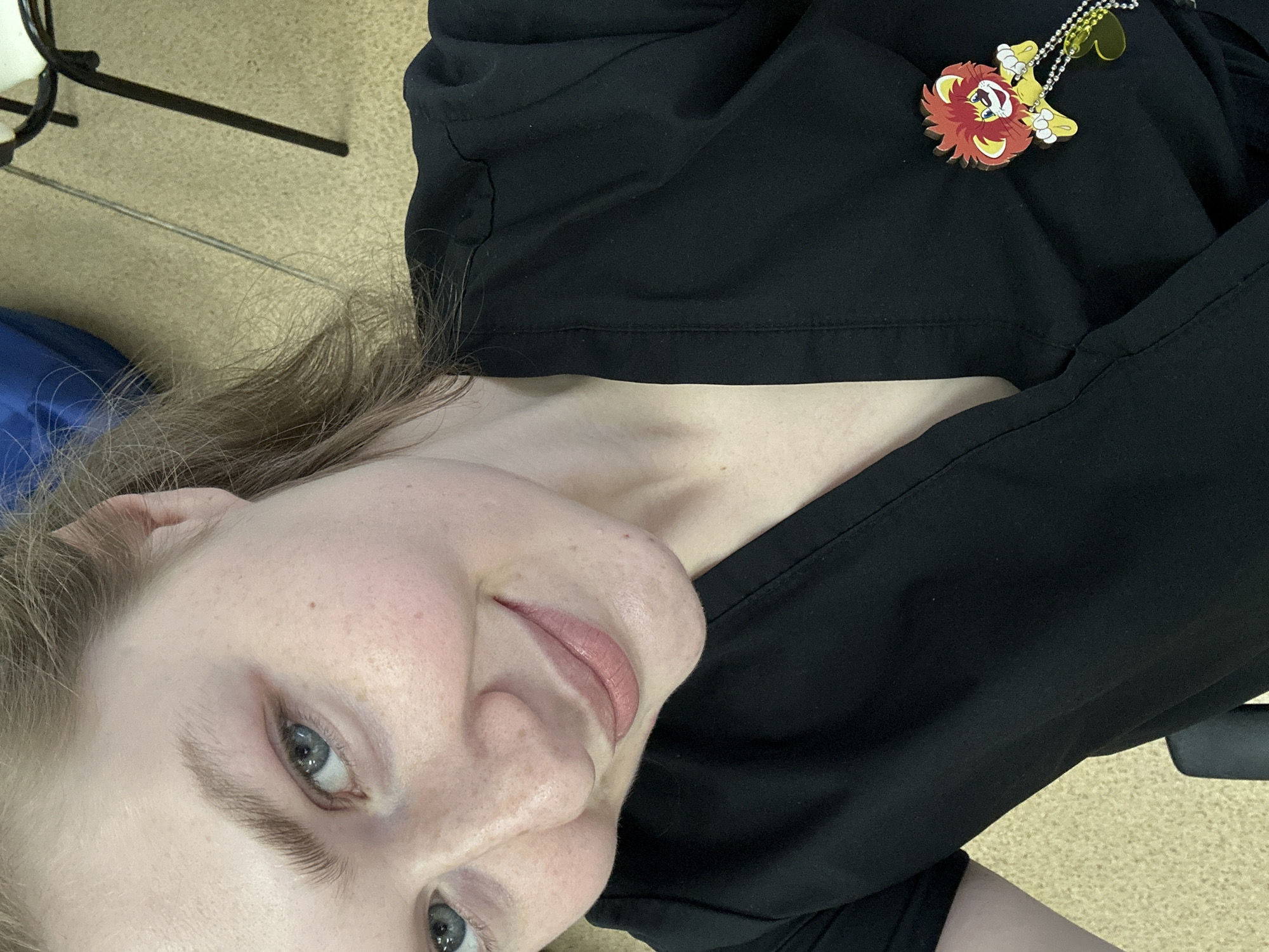 A young woman is depicted in a close-up, chest-up portrait, oriented diagonally in the frame. She has light brown/blonde hair, fair skin with noticeable freckles across her cheeks and nose, and light blue eyes. A subtle, soft smile is present on her lips, conveying a calm or pleasant demeanor.
She is wearing a plain black top, likely scrubs or a similar professional uniform, which covers her shoulders and chest. A vibrant keychain is clipped to her upper right chest area. The keychain features a stylized cartoon lion with a prominent red mane, a yellow face and body, and small white paws, suspended from a small, shiny chain.
The background elements are blurred but suggest an indoor setting. A light-colored, possibly carpeted floor is visible, along with parts of a black metal frame (possibly a chair or stand) and a blue object, which could be part of furniture or a bag. The activity appears to be simply posing for the camera. No discernible text is visible within the image. The setting is an unspecified indoor location, potentially in Kazan, Russia.