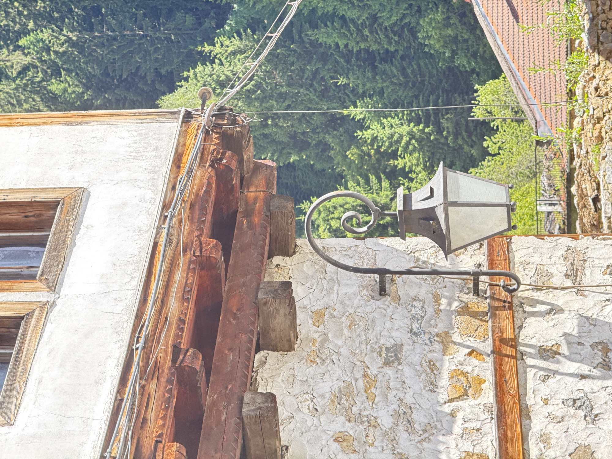 The image shows an exterior wall of a building in Shiroka Laka, Bulgaria. The wall is constructed of rough stone with sections of plaster. A wooden beam runs horizontally across the wall. A black metal lamp hangs from an ornate bracket attached to the wall. The lamp has a glass enclosure. Above the stone wall, there is a wooden eave supported by decorative wooden brackets. Overhead, wires are strung across the scene. In the background, a dense forest of green pine trees is visible. A portion of a red-tiled roof is also present in the lower right corner of the image. The lighting suggests it is daytime.