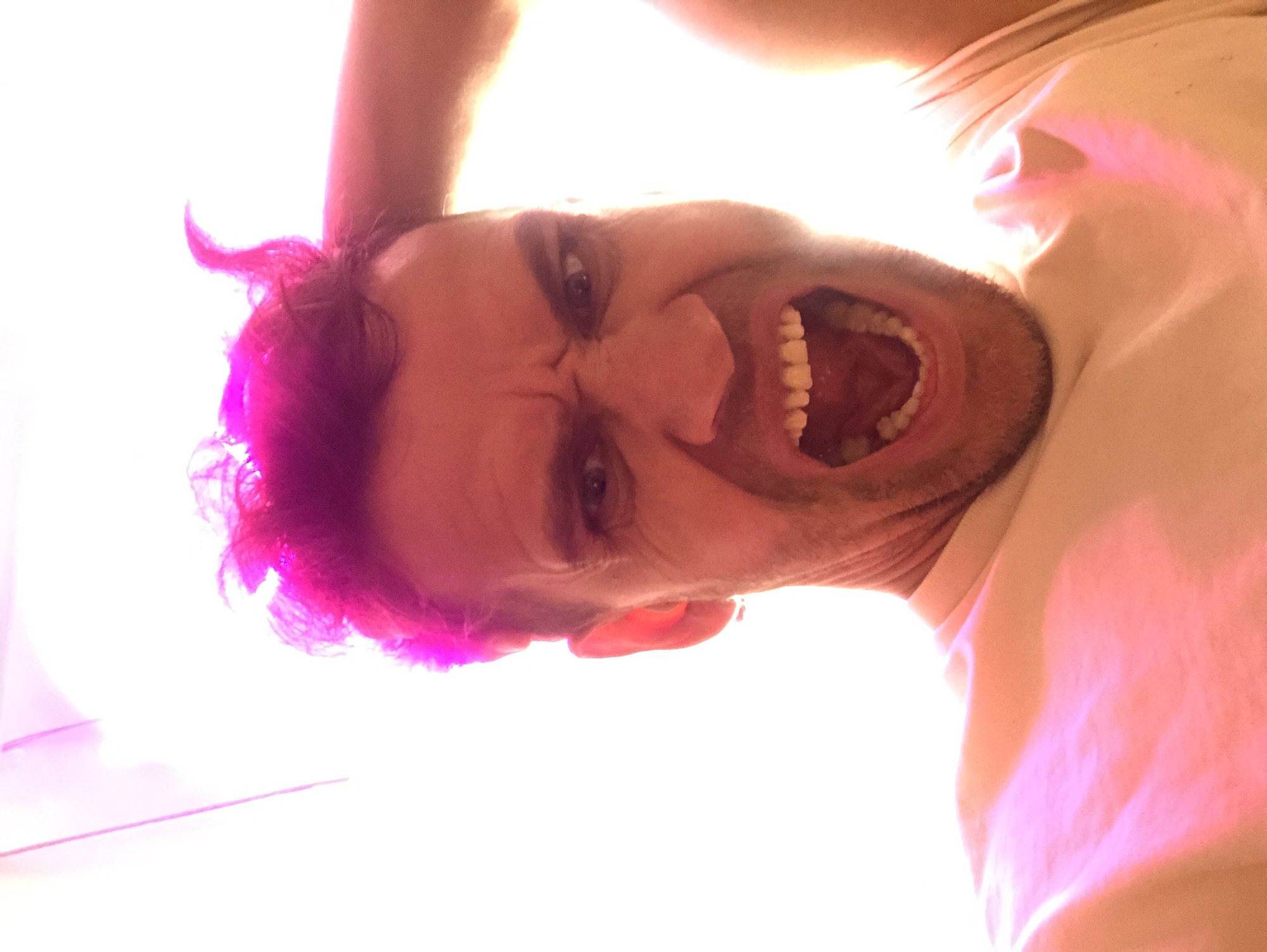 The image shows a close-up, selfie-style shot of a person with short, spiky, bright pink hair. The person's mouth is open in a scream or shout, revealing their teeth. Their eyebrows are furrowed, and their eyes are wide, suggesting an intense emotion. The person's right hand is visible, raised and touching their head. They are wearing a light-colored t-shirt, which has pinkish light reflecting off it. The background is very bright and appears to be a white or light-colored wall or ceiling, with a faint pinkish-purple light source visible in the upper left corner. There is no discernible location in the city of Saint Petersburg, Russia, or any environmental details to indicate such.