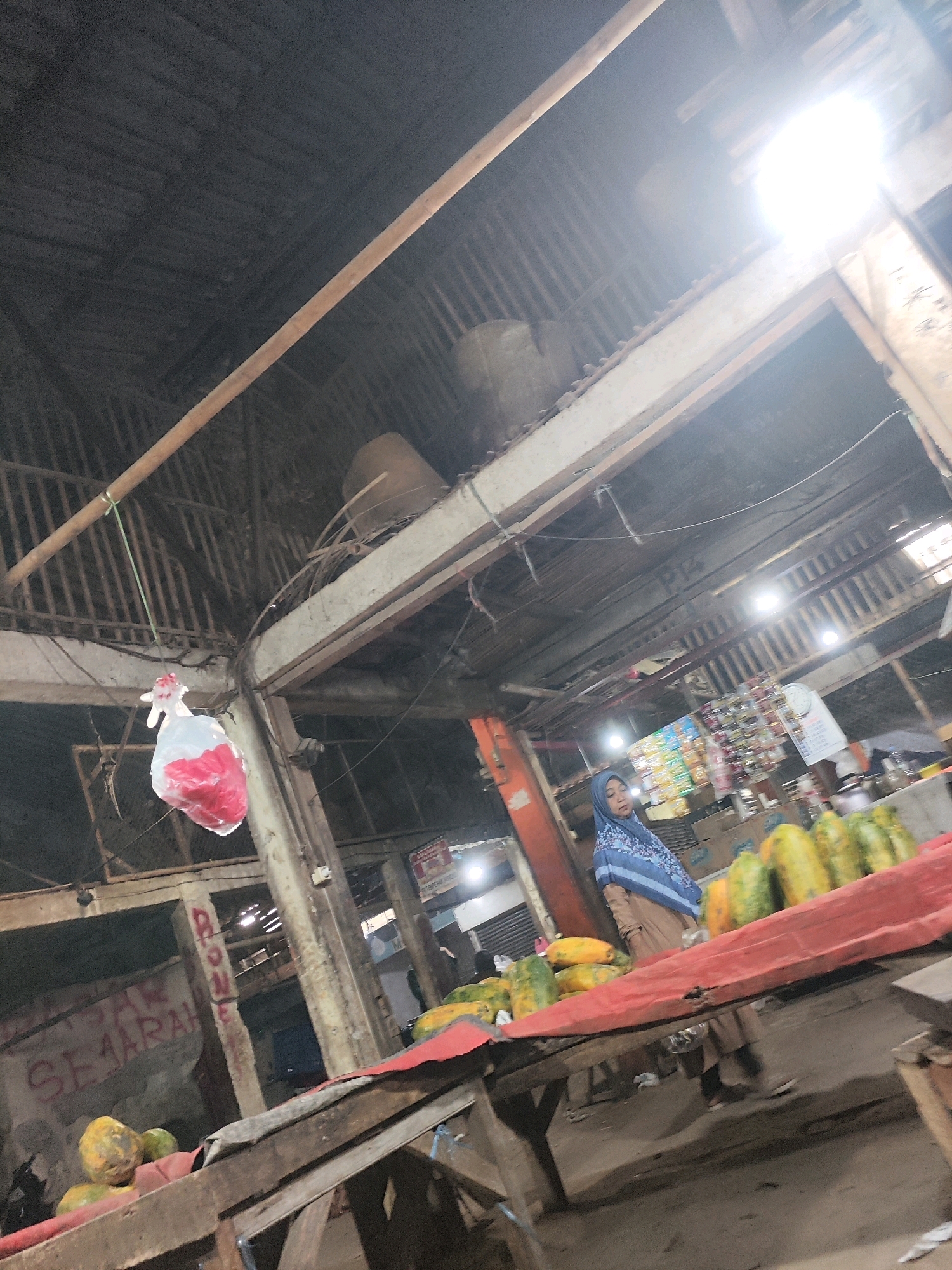 The image depicts an indoor market scene, likely in Karawang, Indonesia, characterized by a rustic, open-air structure with a corrugated metal roof and wooden beams. In the foreground, a wooden stall is laden with a variety of fruits, including papayas. A woman wearing a blue hijab and a brown abaya stands behind the stall, appearing to be a vendor. To the left, a white plastic bag filled with a red substance hangs from a string. The background reveals more stalls with various goods, including packaged items and a calendar. The overall atmosphere is that of a busy, local marketplace.